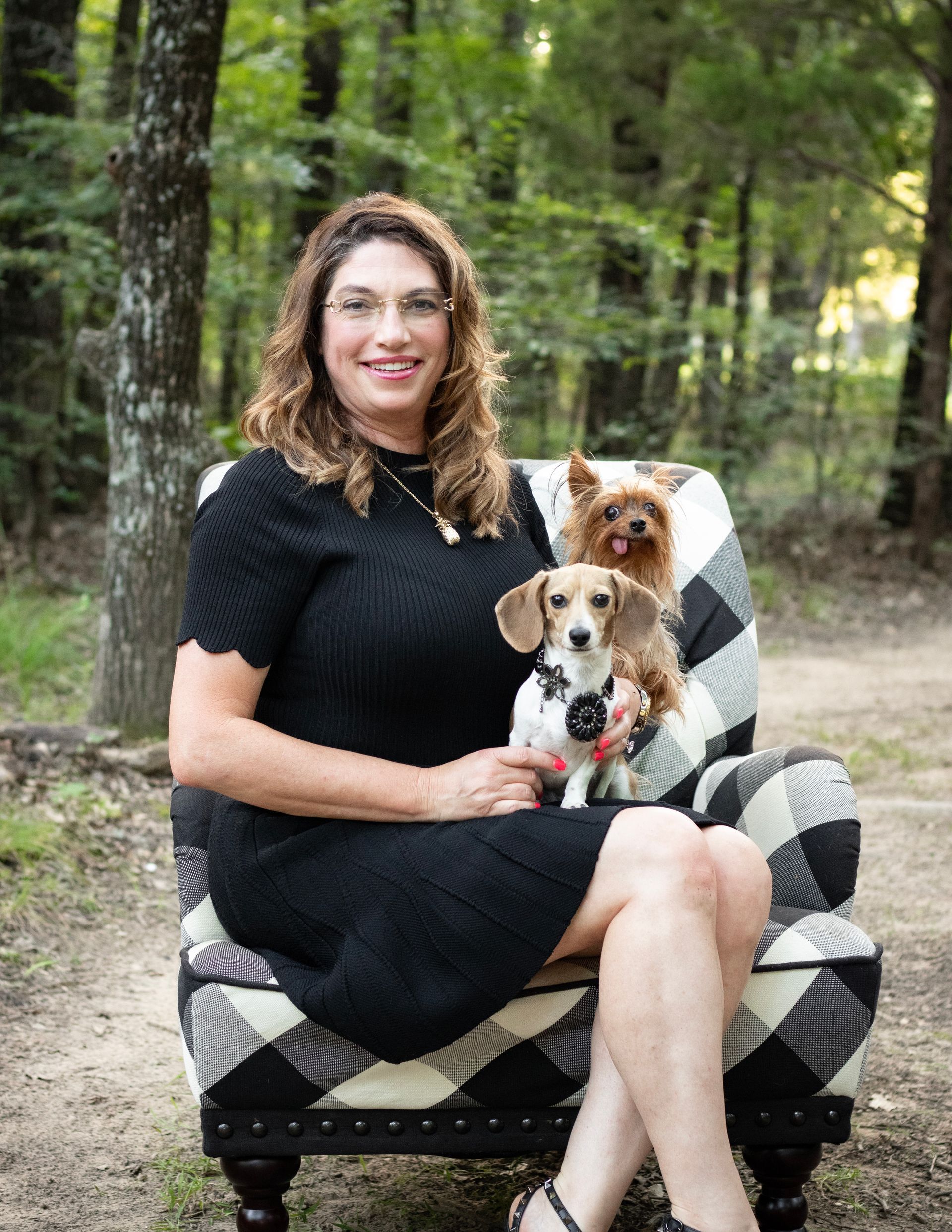 Woman in black dress sits with two small dogs on a checkered armchair in a forest setting.