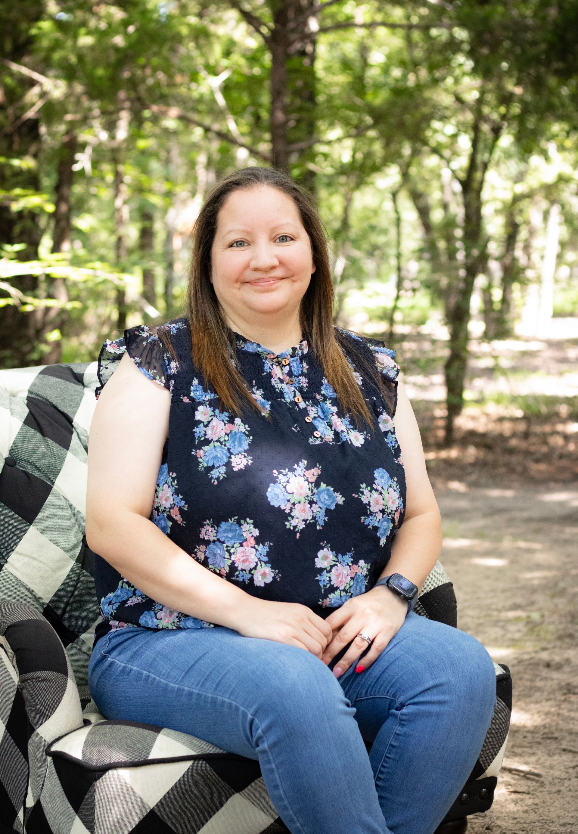 Woman in floral top and jeans sits on a plaid chair outdoors, smiling.