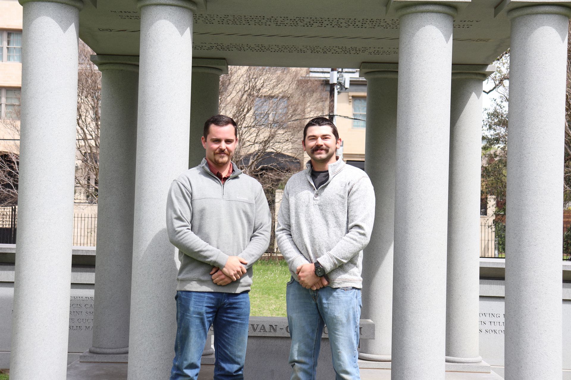 Two men stand in front of stone columns, both wearing gray sweaters and jeans, smiling.