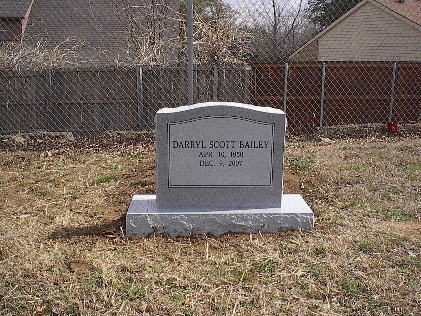 Gray headstone on a gray base, inscribed with a name and dates, in a grassy yard behind a chain-link fence.