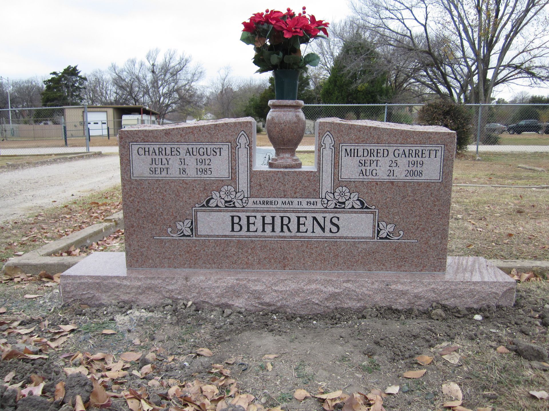 Headstone for the Behrens family with names and dates, topped with a flower arrangement.