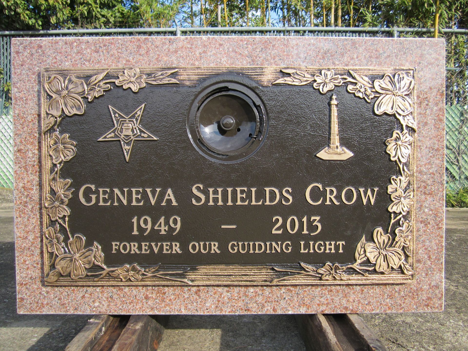 Headstone for Geneva Shields Crow, bronze plaque on pink granite. Contains a star, lighthouse, speaker, and floral design.