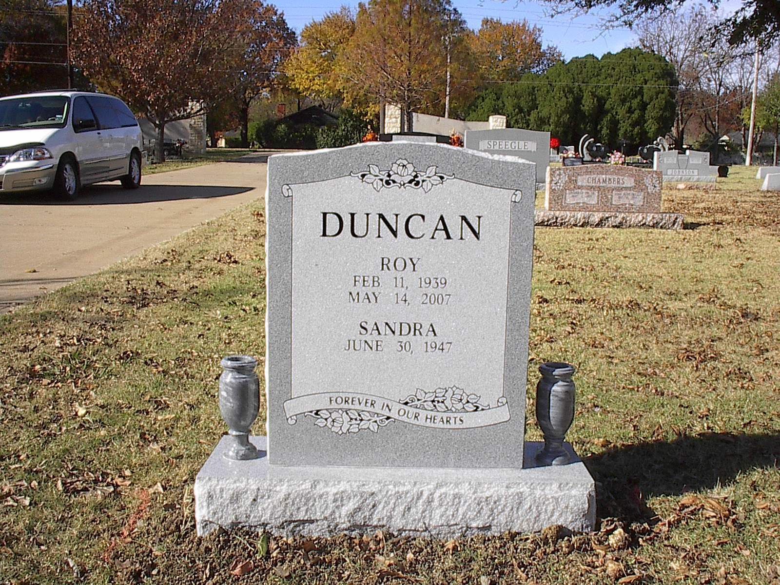 Gravestone of Roy and Sandra Duncan in a cemetery, with a gray vehicle in the background.