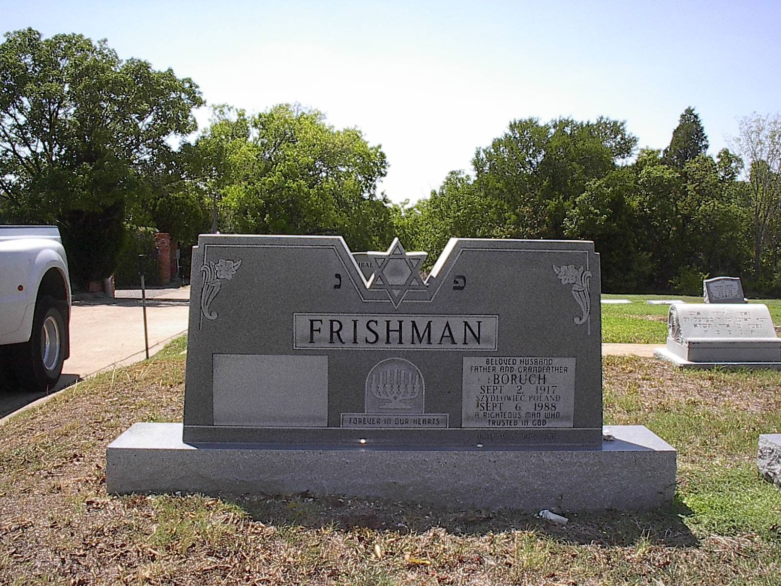 Gray Frishman family headstone in a cemetery with a white car in the background.