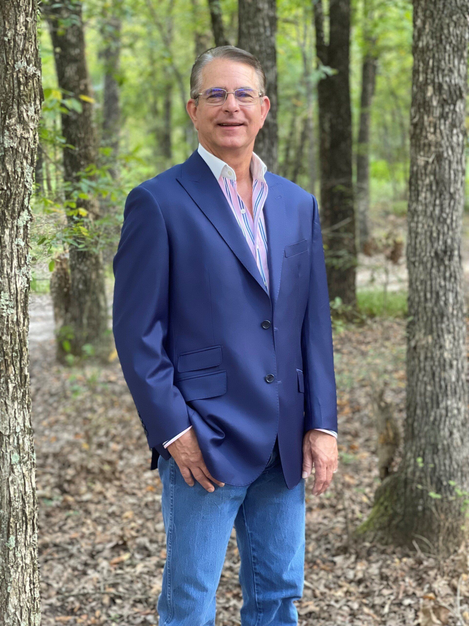 Man in blue blazer, patterned shirt, and jeans stands in a wooded area.