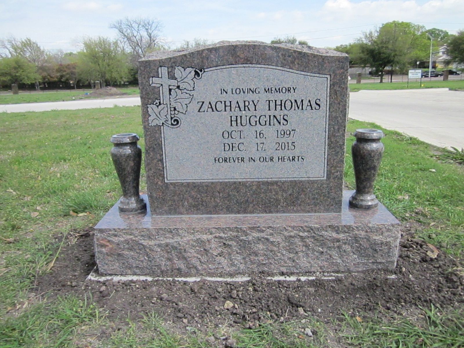 Grave marker for Zachary Thomas Huggins, with cross and leaves design, in a grassy cemetery.