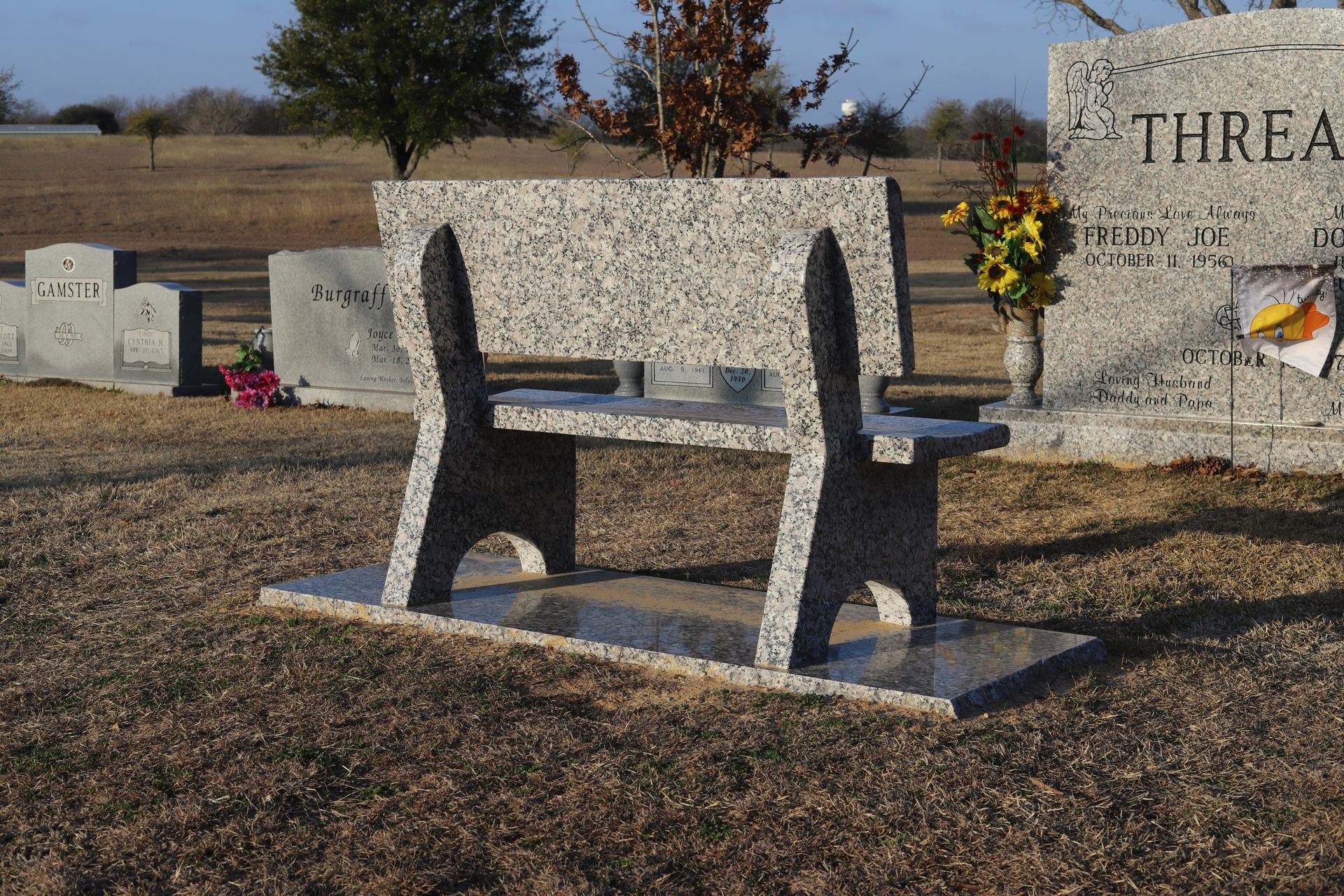 Granite bench in a cemetery with headstones and dried grass.