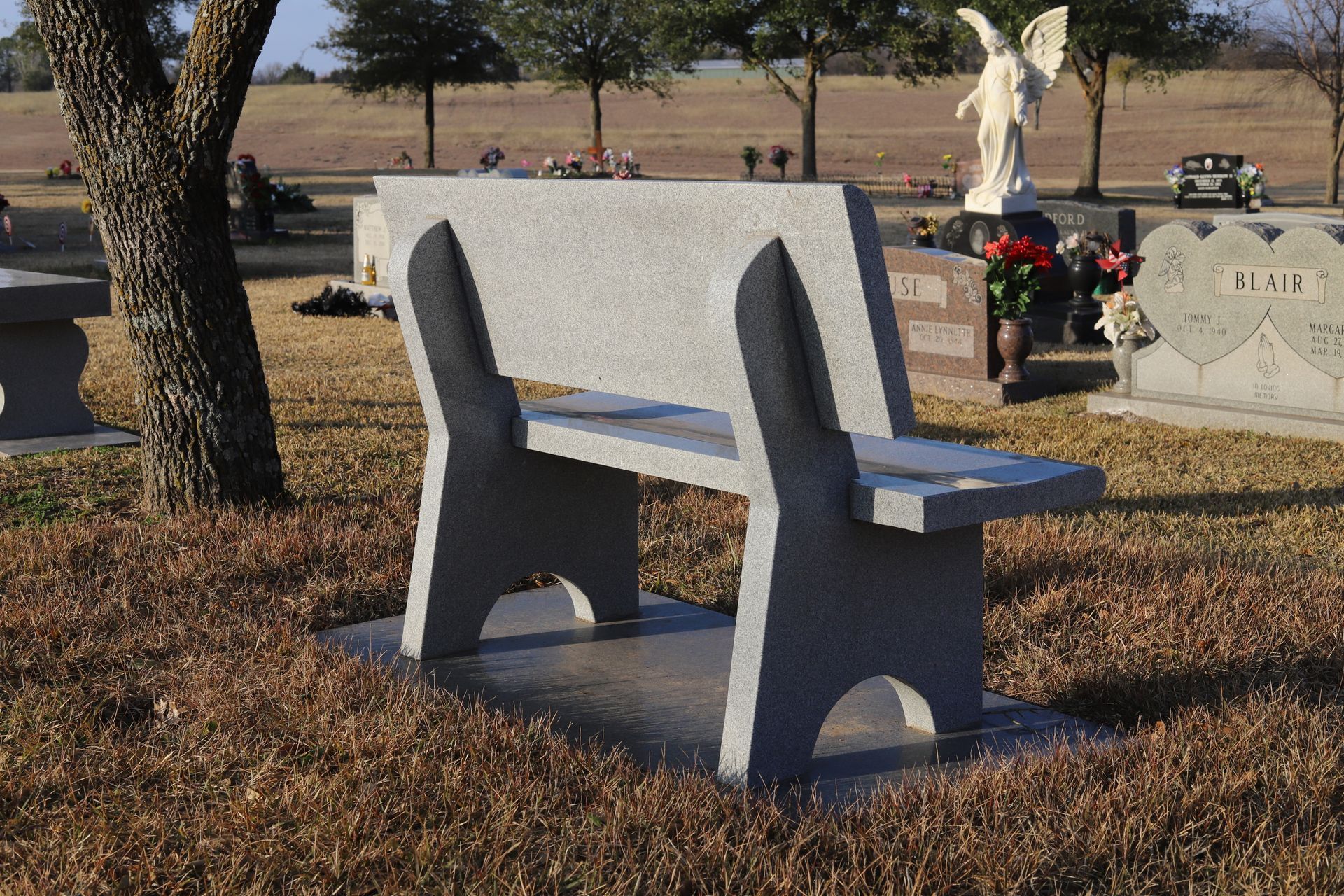 Stone bench in a cemetery with a statue of an angel and tombstones in the background.