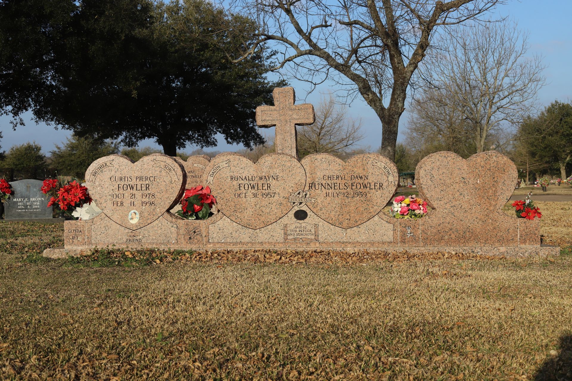 Grave marker with heart-shaped headstones, cross, names, and flowers in a grassy cemetery.