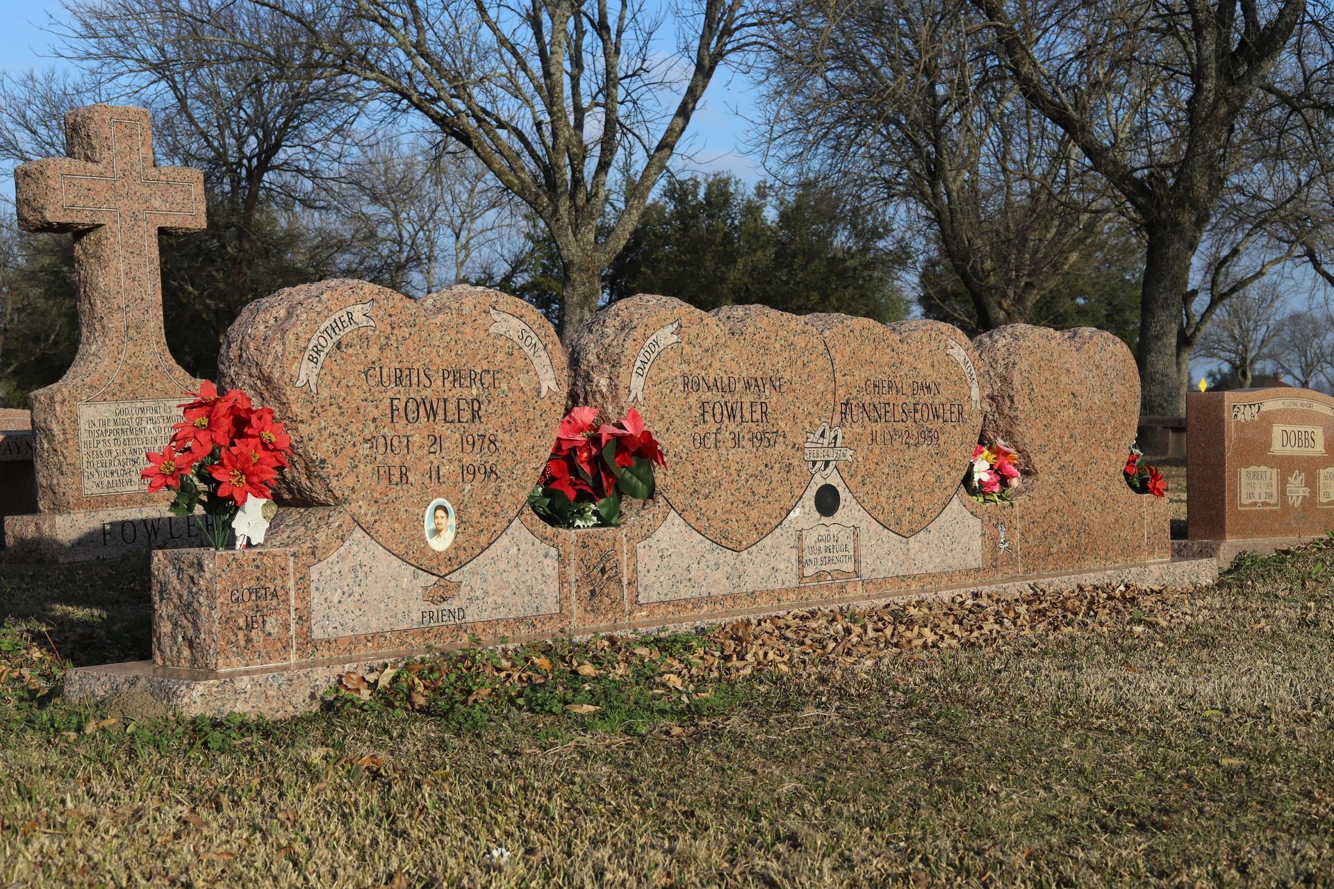 Heart-shaped tombstones with flower arrangements in a grassy cemetery. A cross stands nearby.