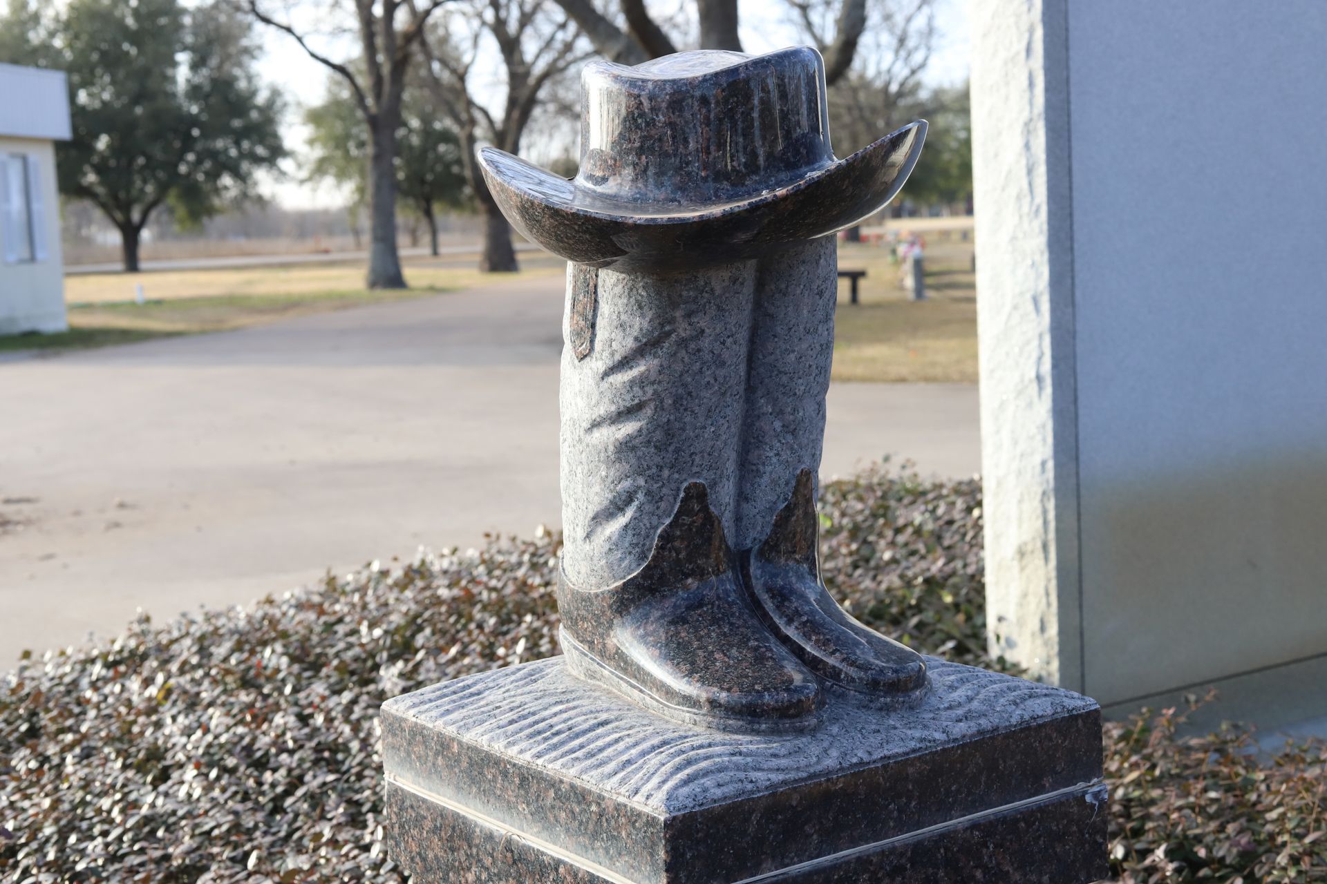 Stone sculpture of cowboy boots and hat on a pedestal, outdoors in a park-like setting.