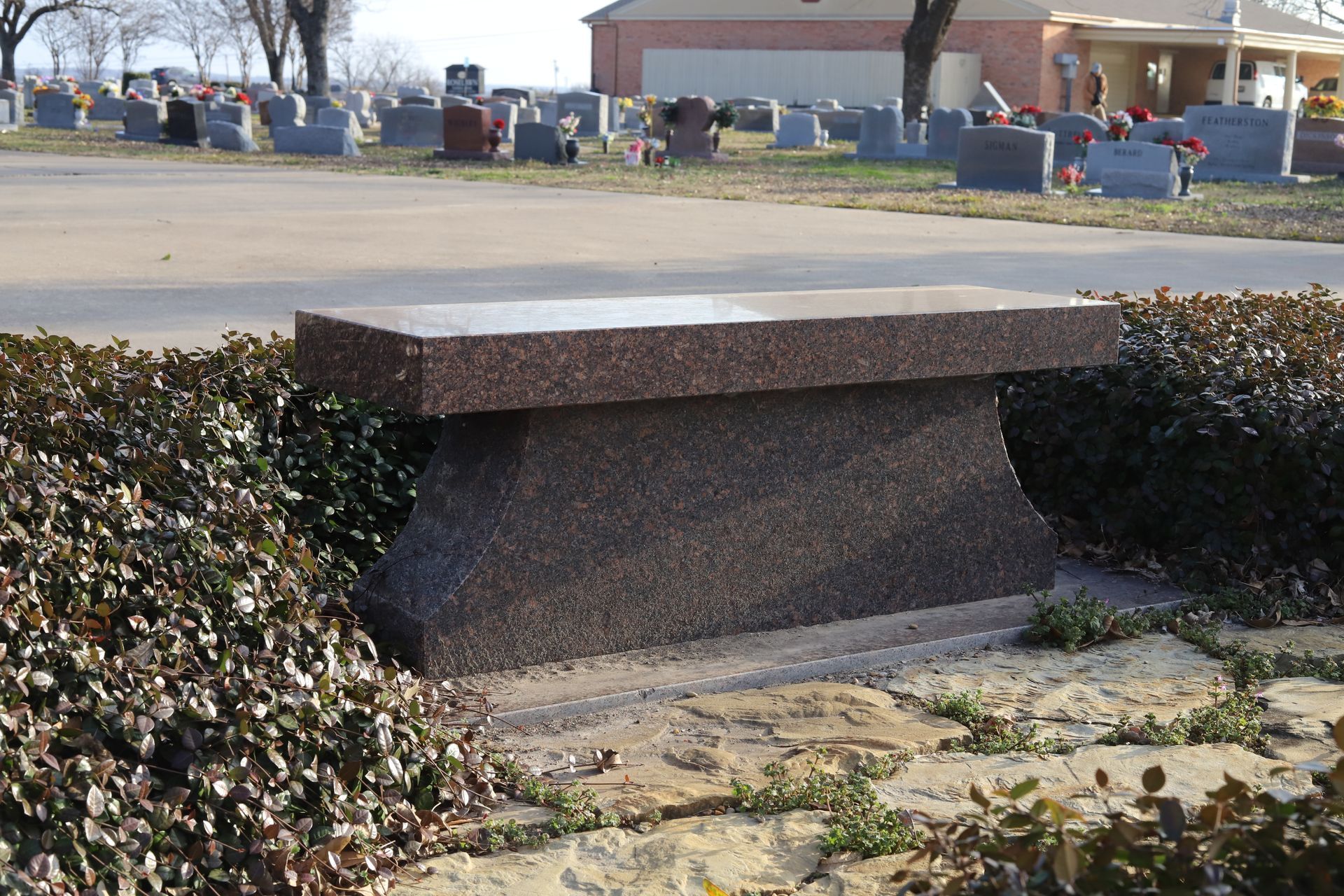 Granite bench in a cemetery, surrounded by bushes. Graves and a building are in the background.