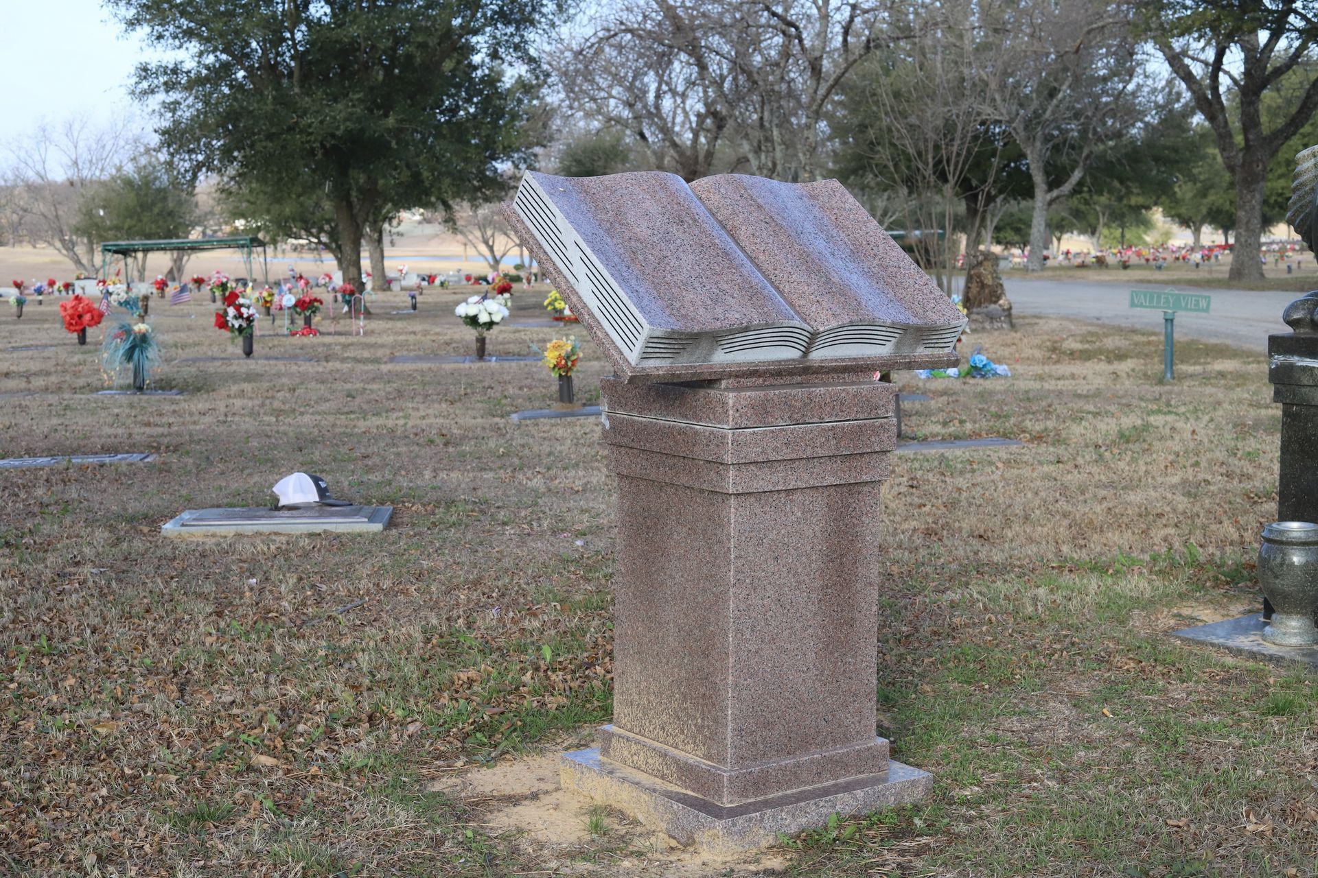 A cemetery with a large, open book-shaped headstone on a pedestal. Rows of other headstones and trees are in the background.