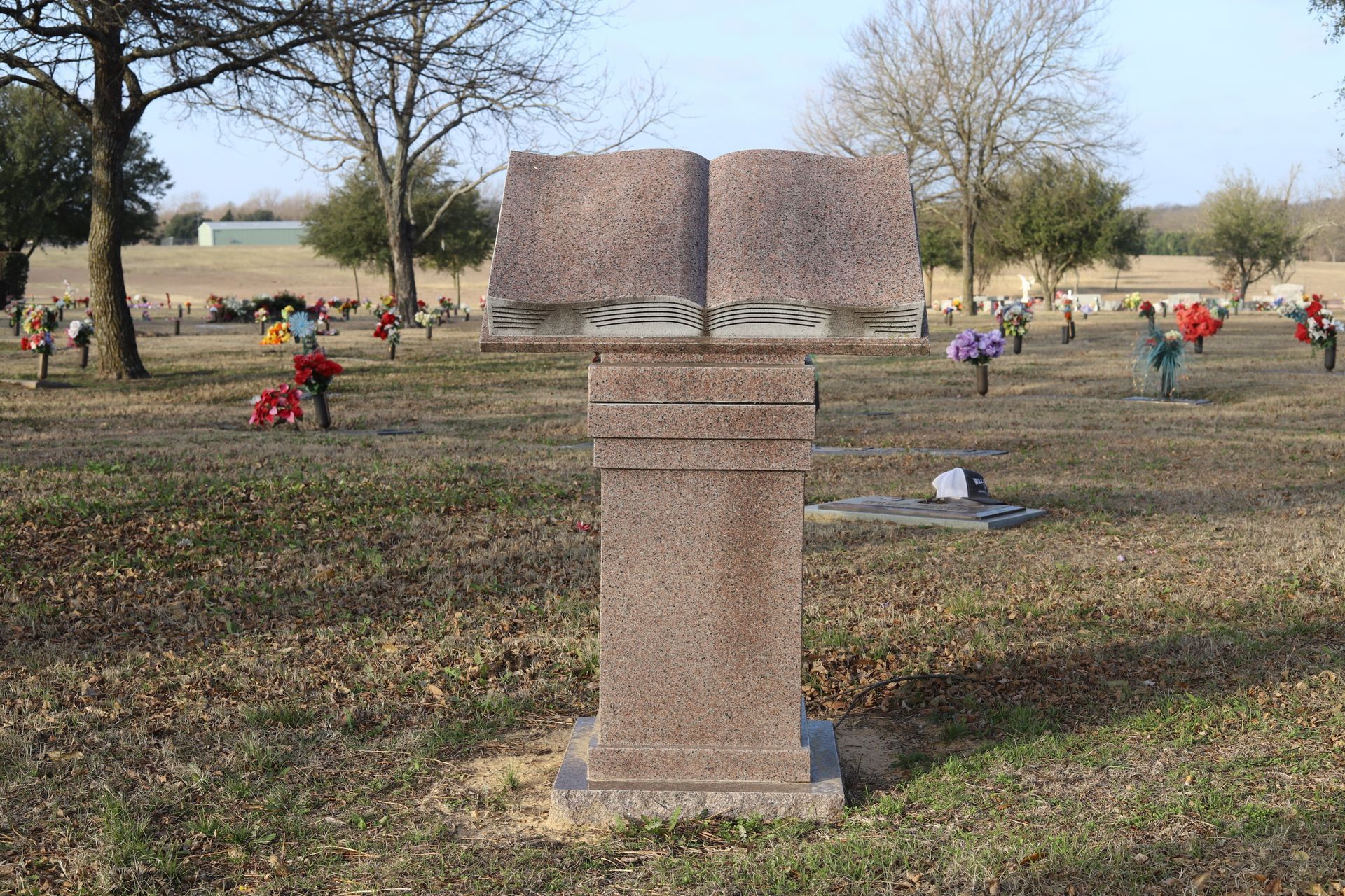 Stone monument in a cemetery shaped like an open book; other headstones and flowers in the background.