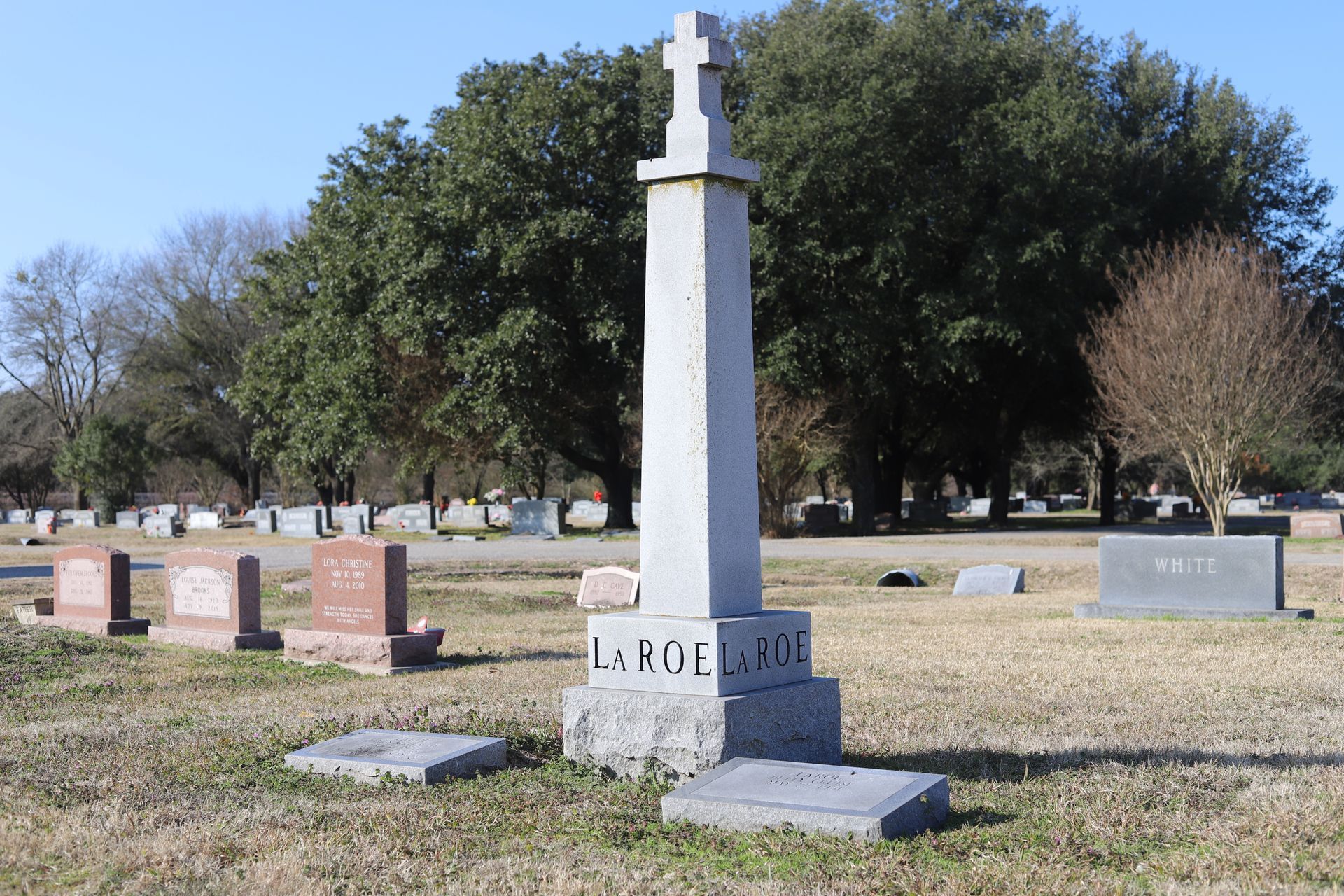 Tall stone grave marker with cross at the top in a cemetery, surrounded by other headstones on a sunny day.