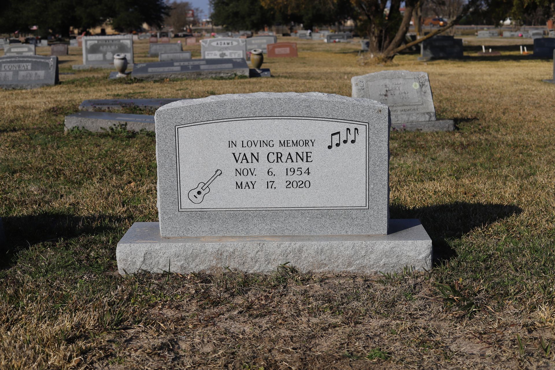 Headstone for Van Crane in a cemetery. Gray granite with musical notes and dates: May 6, 1954 - May 11, 2020.