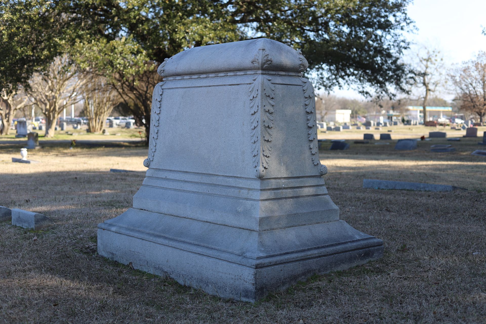 Gray stone tombstone in a cemetery; it has a rectangular base and decorative side details.