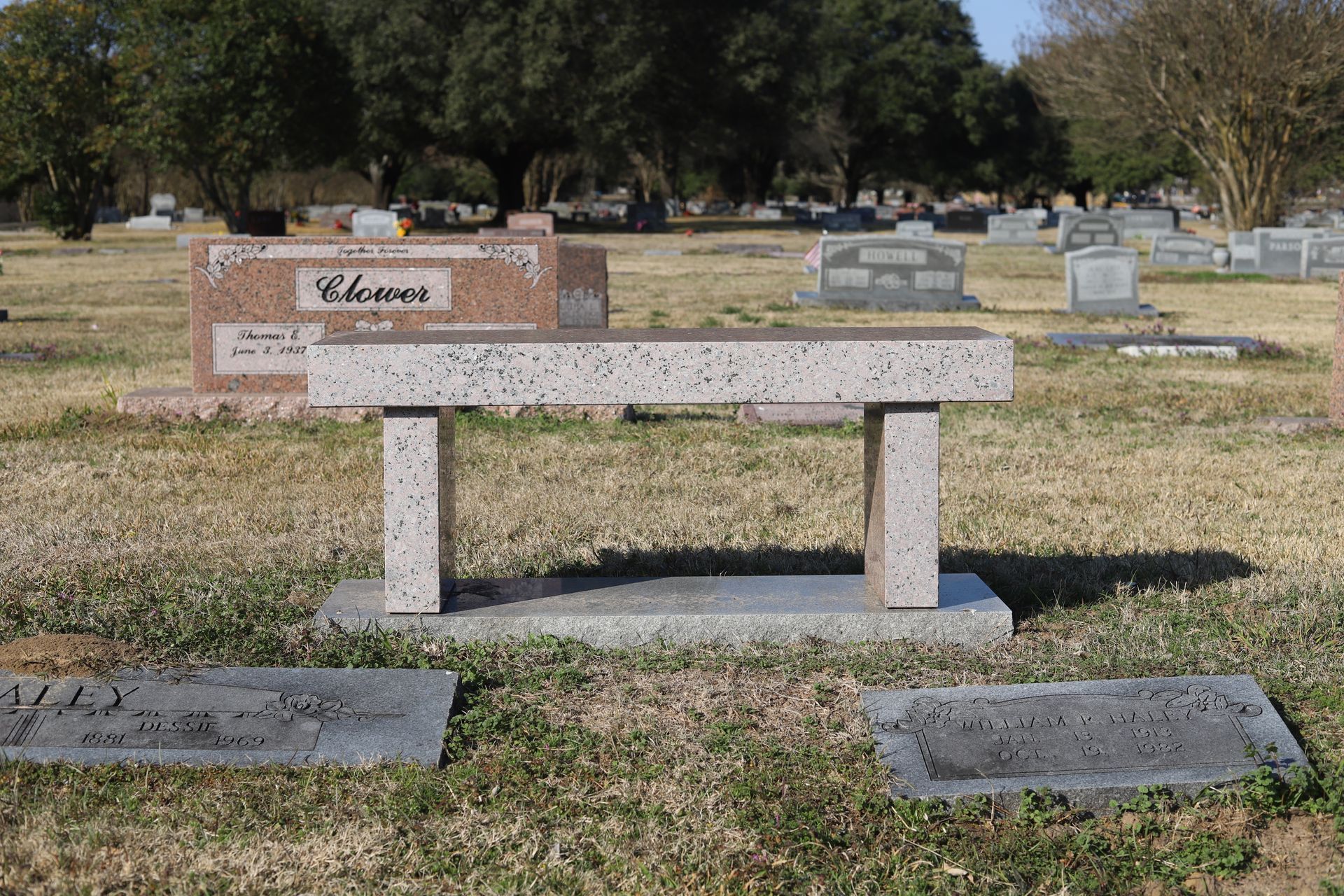 A granite bench in a cemetery, with tombstones in the background under a blue sky.