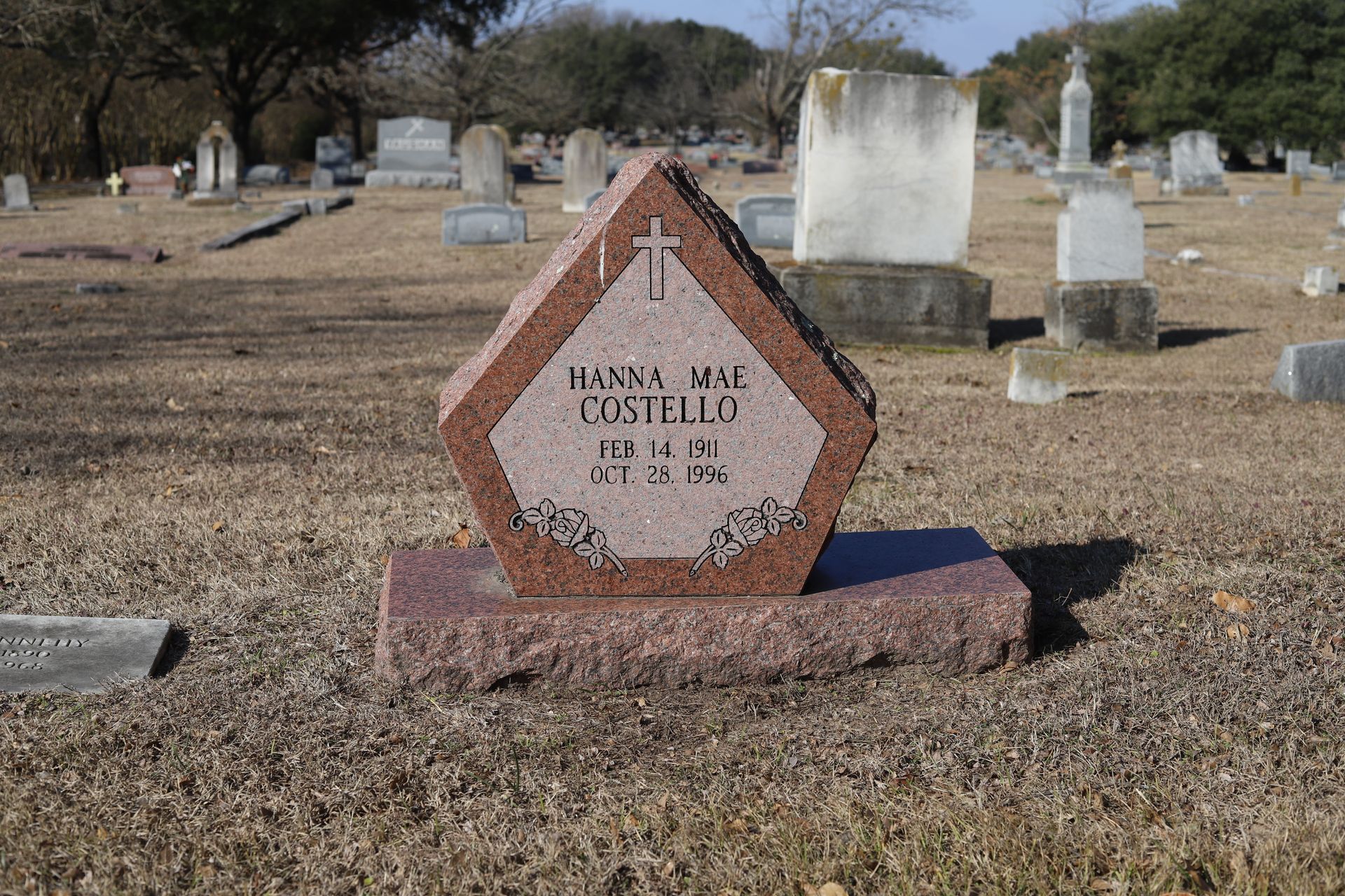 Red granite headstone with name, dates, and cross in a cemetery setting.