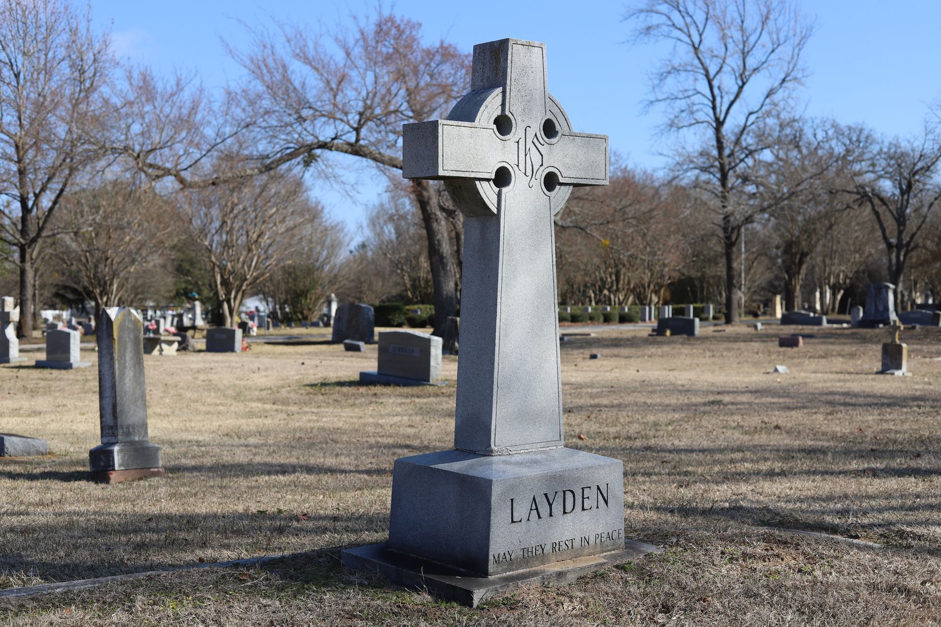 A Celtic cross gravestone in a cemetery with other markers, under a blue sky.
