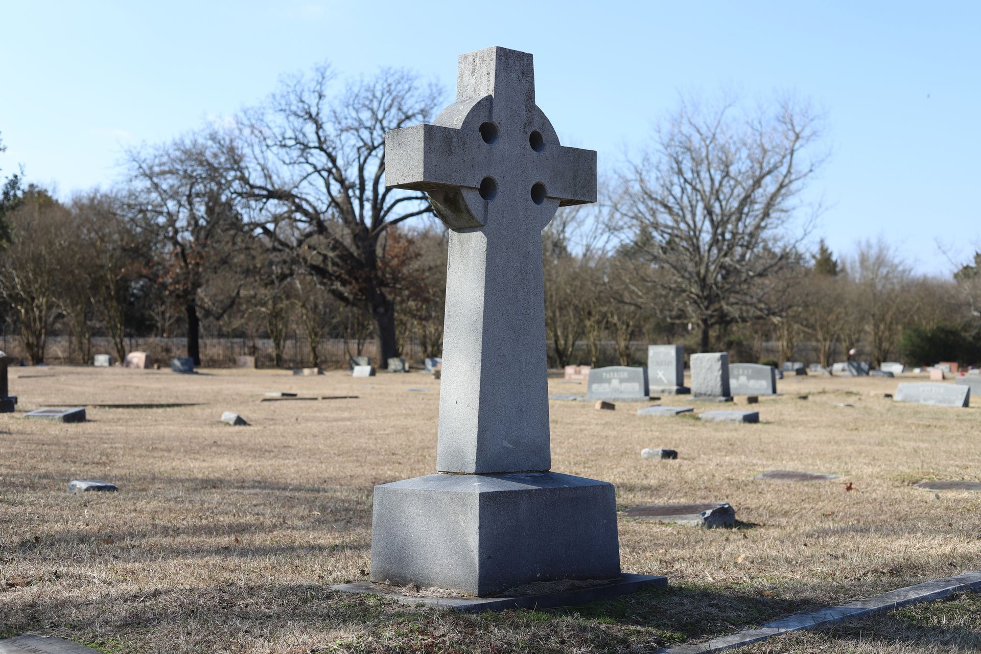 Celtic cross tombstone in a cemetery with other markers and trees.