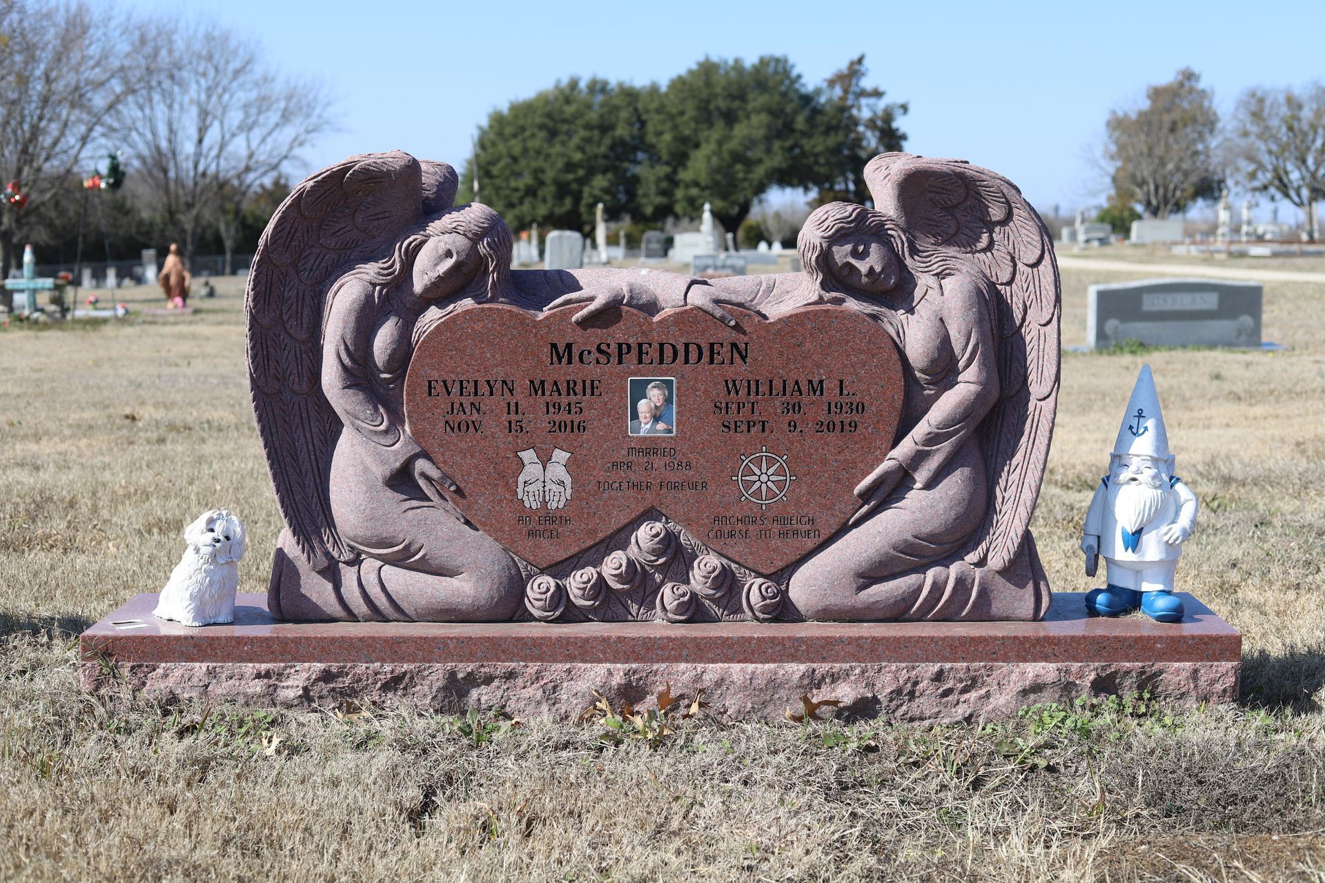 Pink granite headstone in a cemetery, featuring two angels, holding a heart with inscriptions and photos.