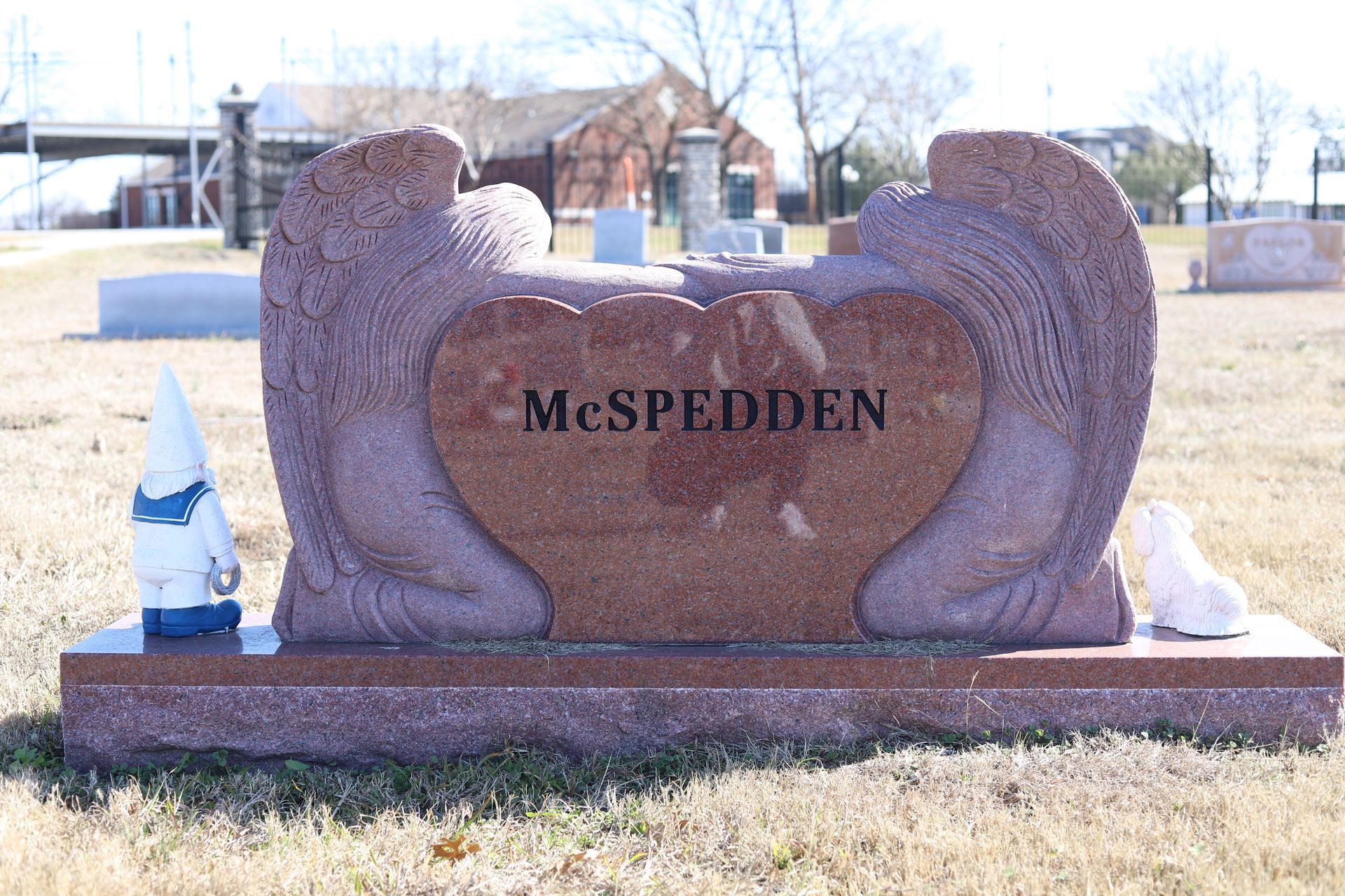 Pink granite McSpedden tombstone with angel wings, a heart, and a gnome statue in a cemetery.