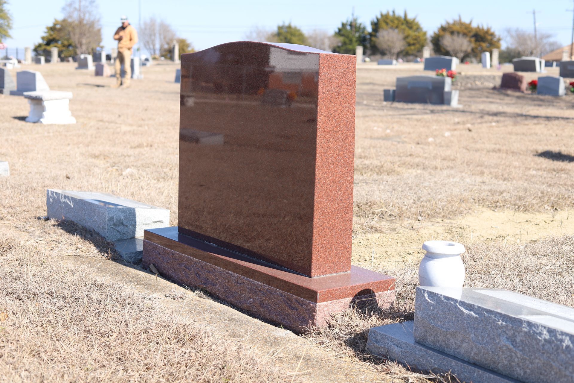 Red granite headstone in a cemetery on a sunny day. A person walks in the background.