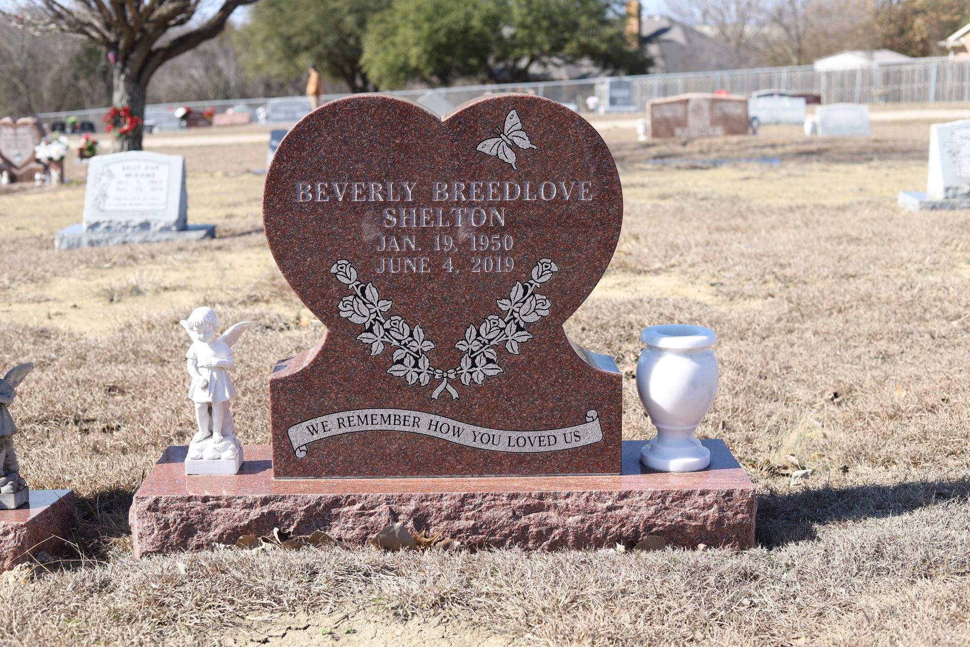 Red heart-shaped tombstone with a butterfly, flowers, and a vase, in a cemetery.