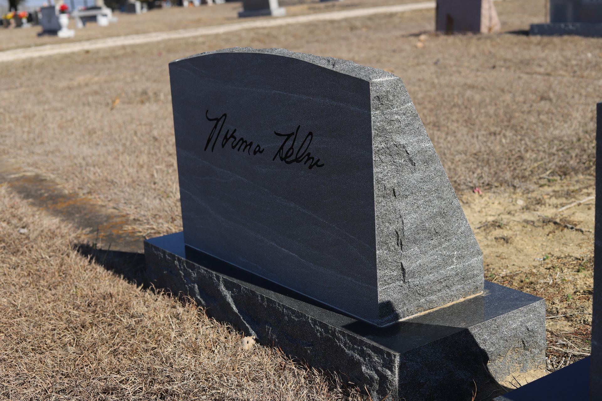 Grave marker in a cemetery; gray granite, engraved with a signature, set on a stone base, brown grass in the background.