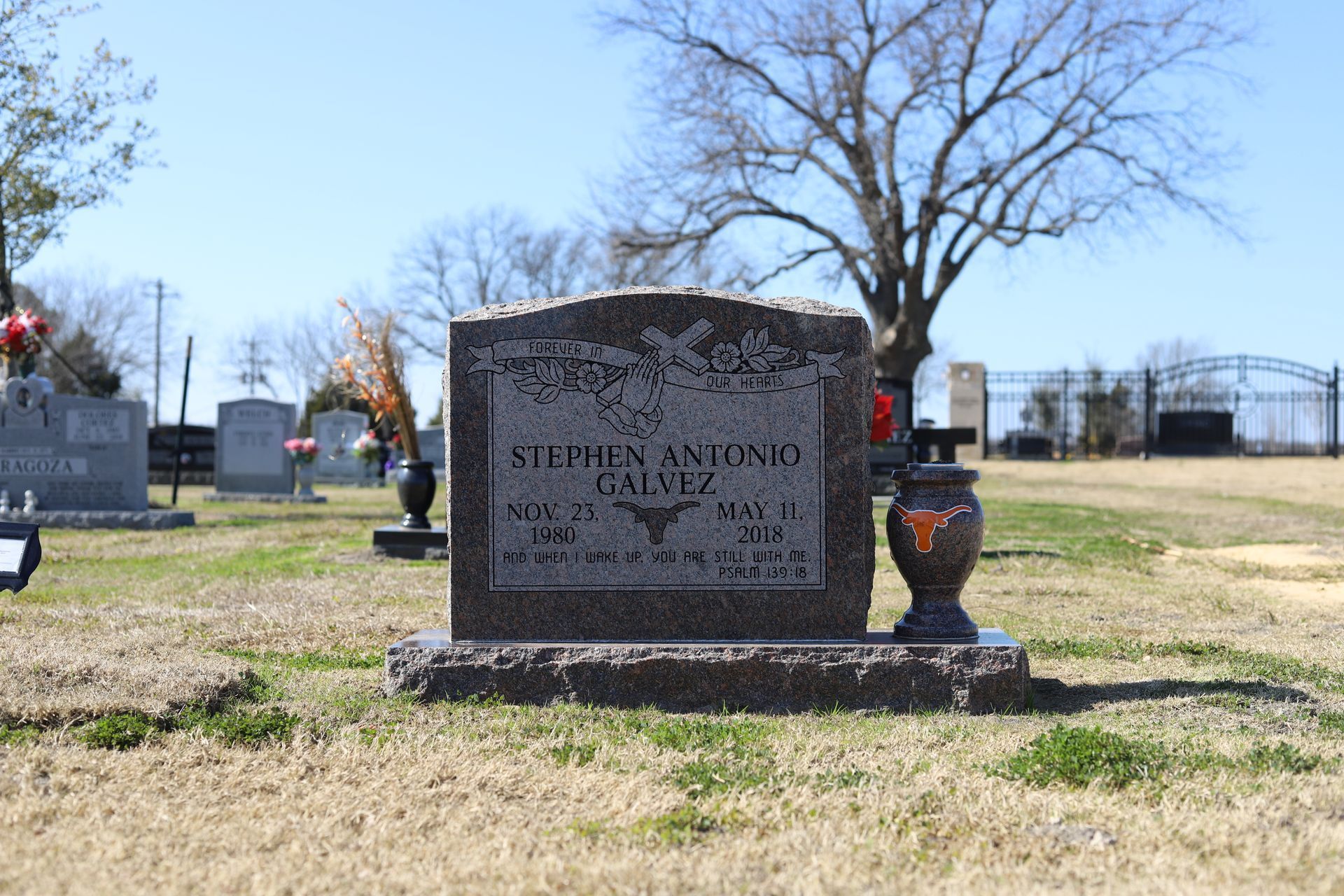 Gravestone in a cemetery, with etched name and dates, Texas Longhorns emblem, vase on the right.