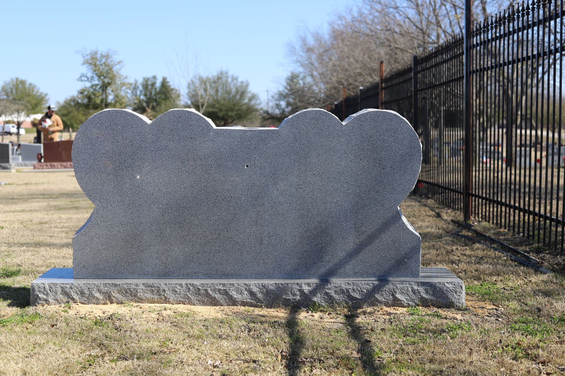Gray headstone with rounded shapes at the top, in a cemetery with a black fence.