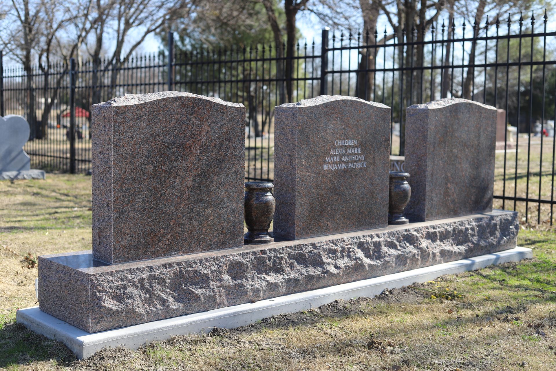 Headstones in a cemetery; three granite monuments with inscriptions, vases on a stone base, black fence in the background.