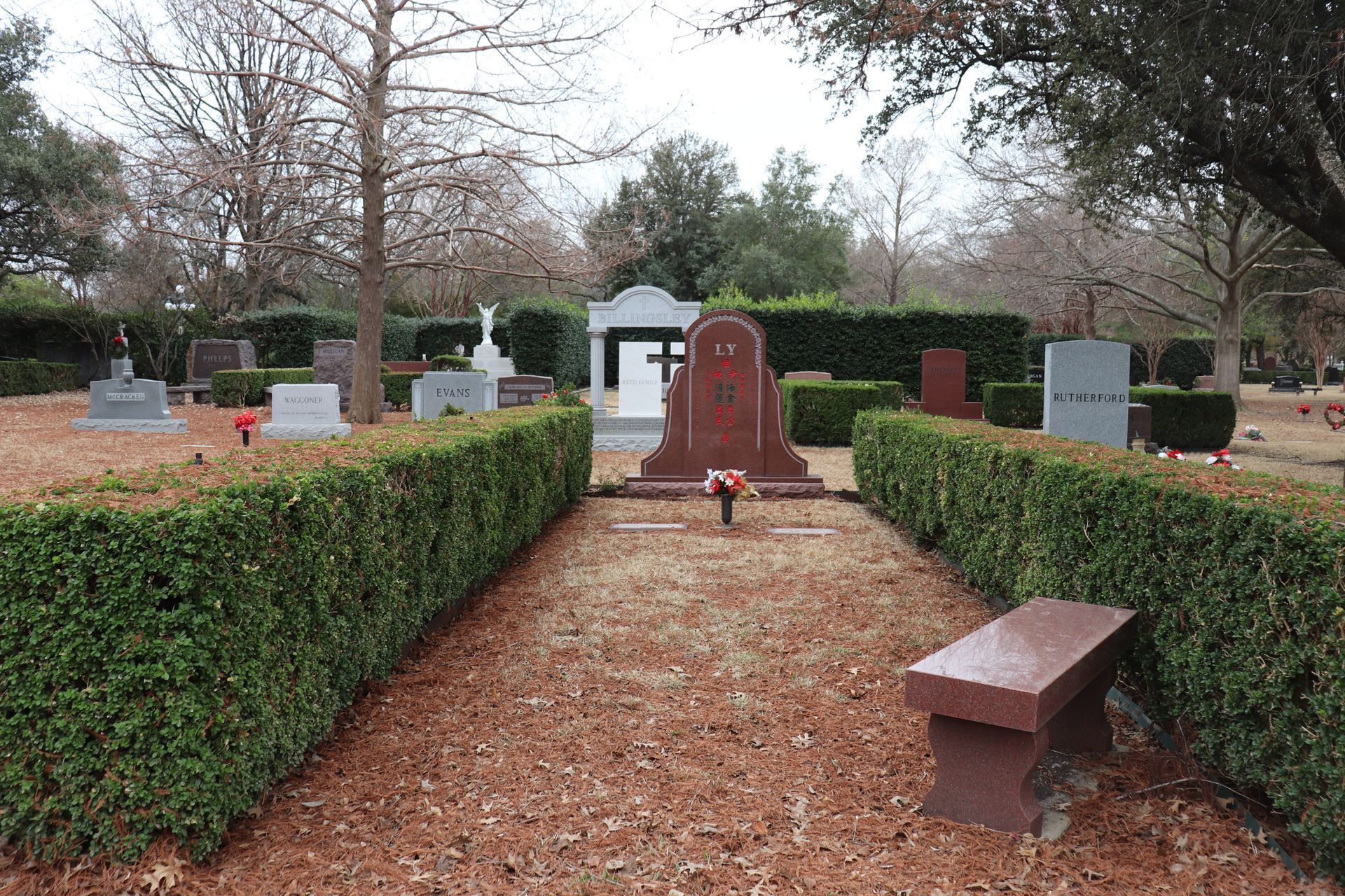 Cemetery scene with headstones, a bench, and hedges. Overcast sky. Brown ground cover.
