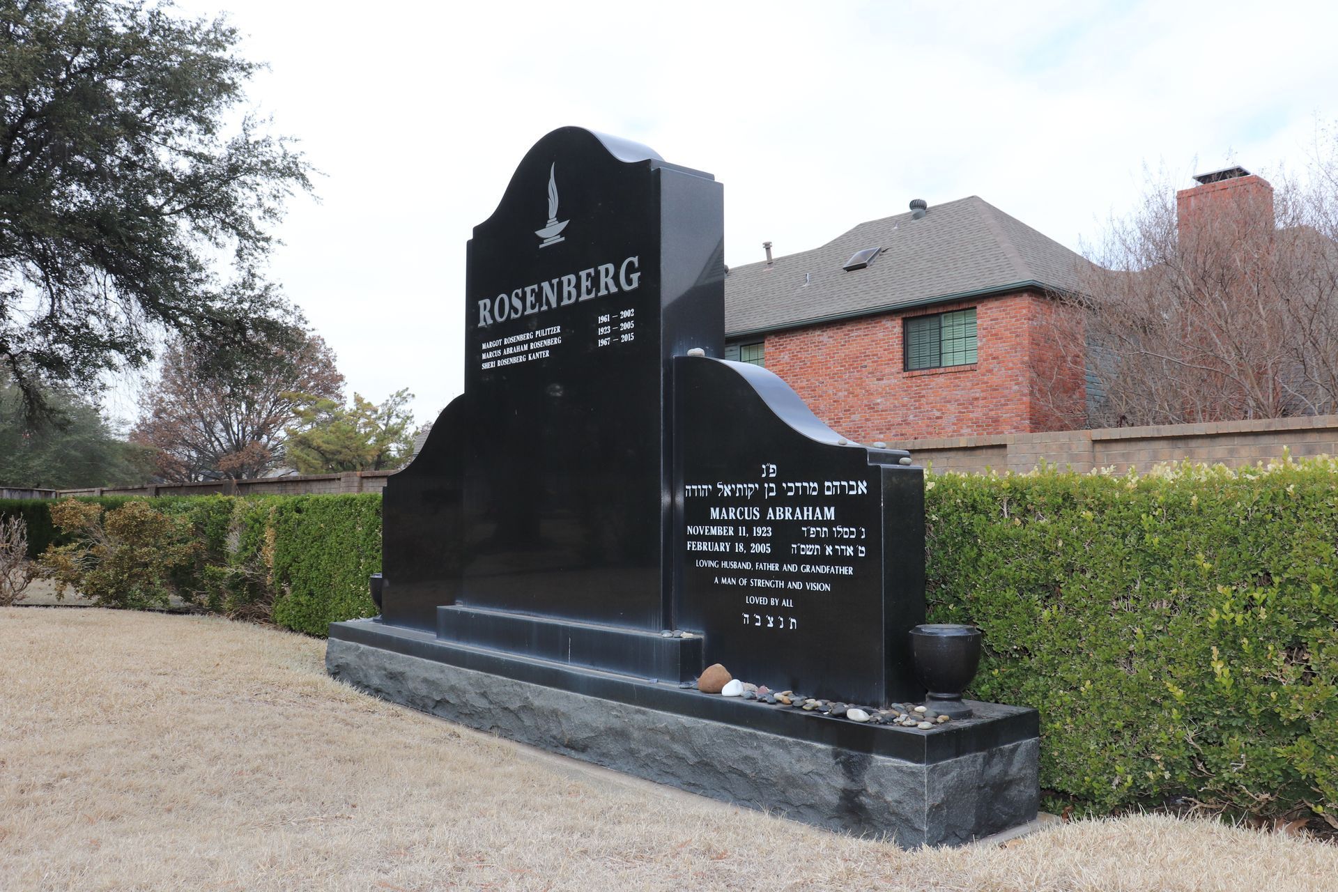 Black headstone in a cemetery with Hebrew writing and a tall, arched top; a house and hedge are in the background.