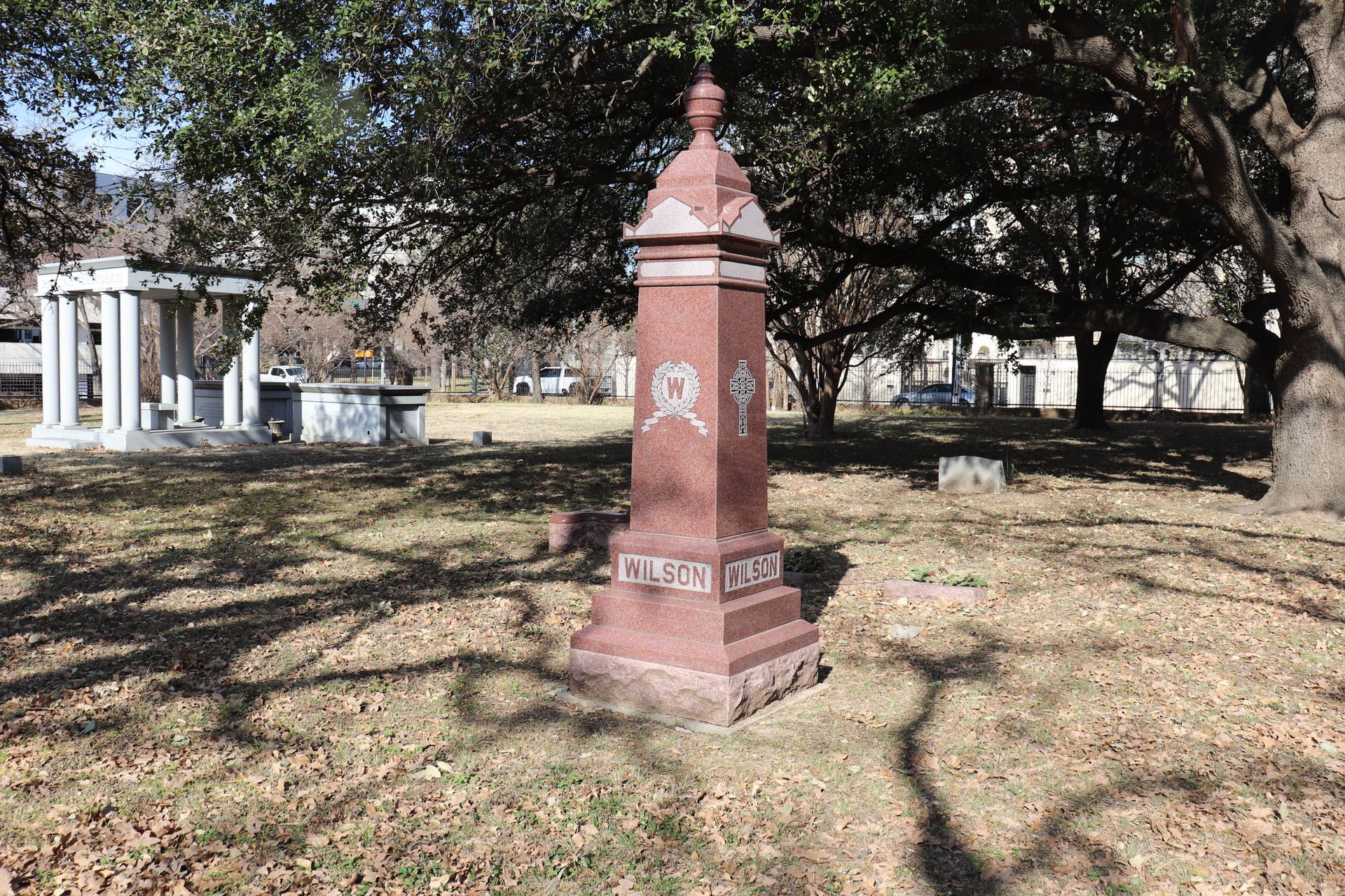Red granite tombstone in a cemetery with a mausoleum in the background on a sunny day.