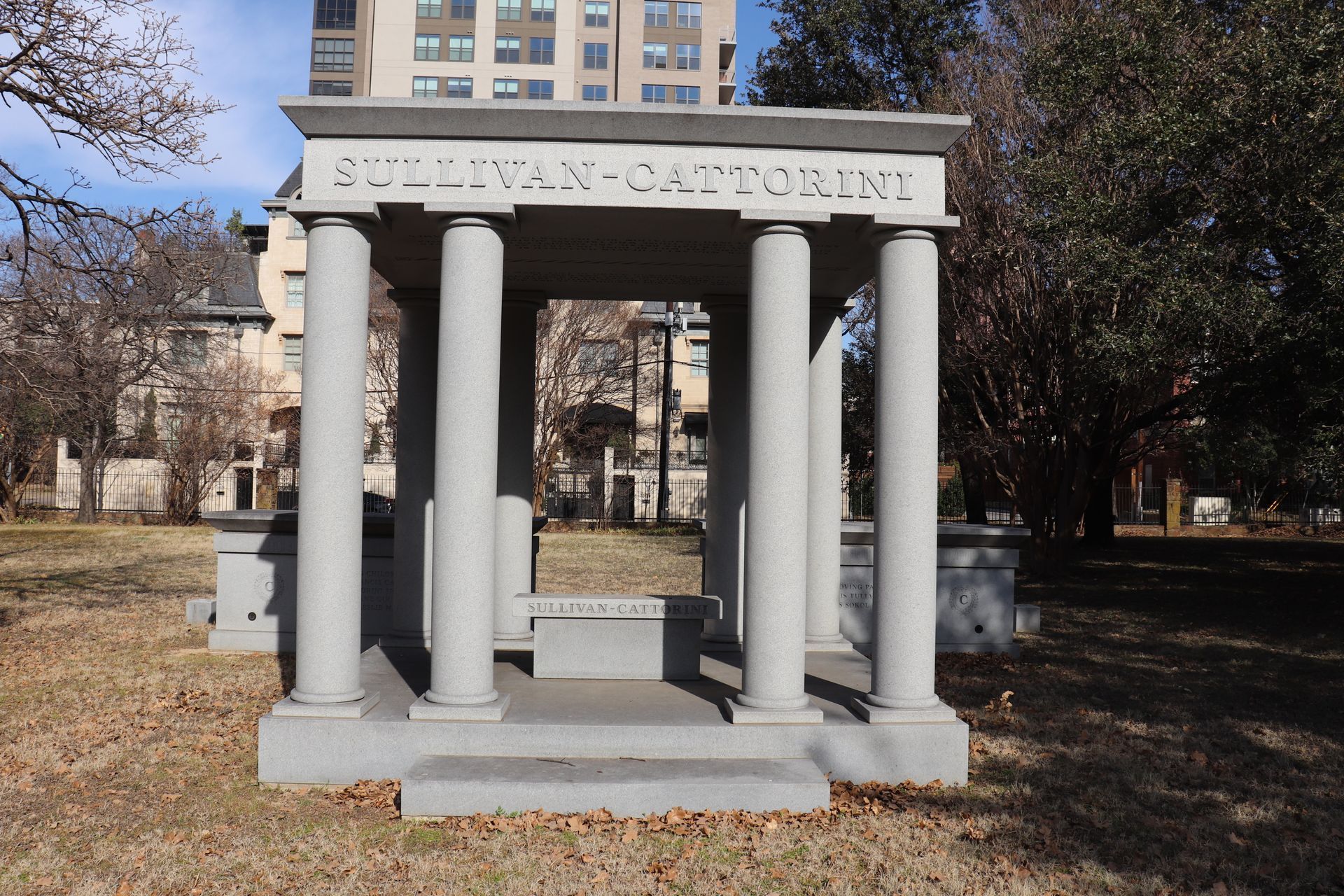 Gray stone memorial structure with columns, in a grassy cemetery. The inscription reads 
