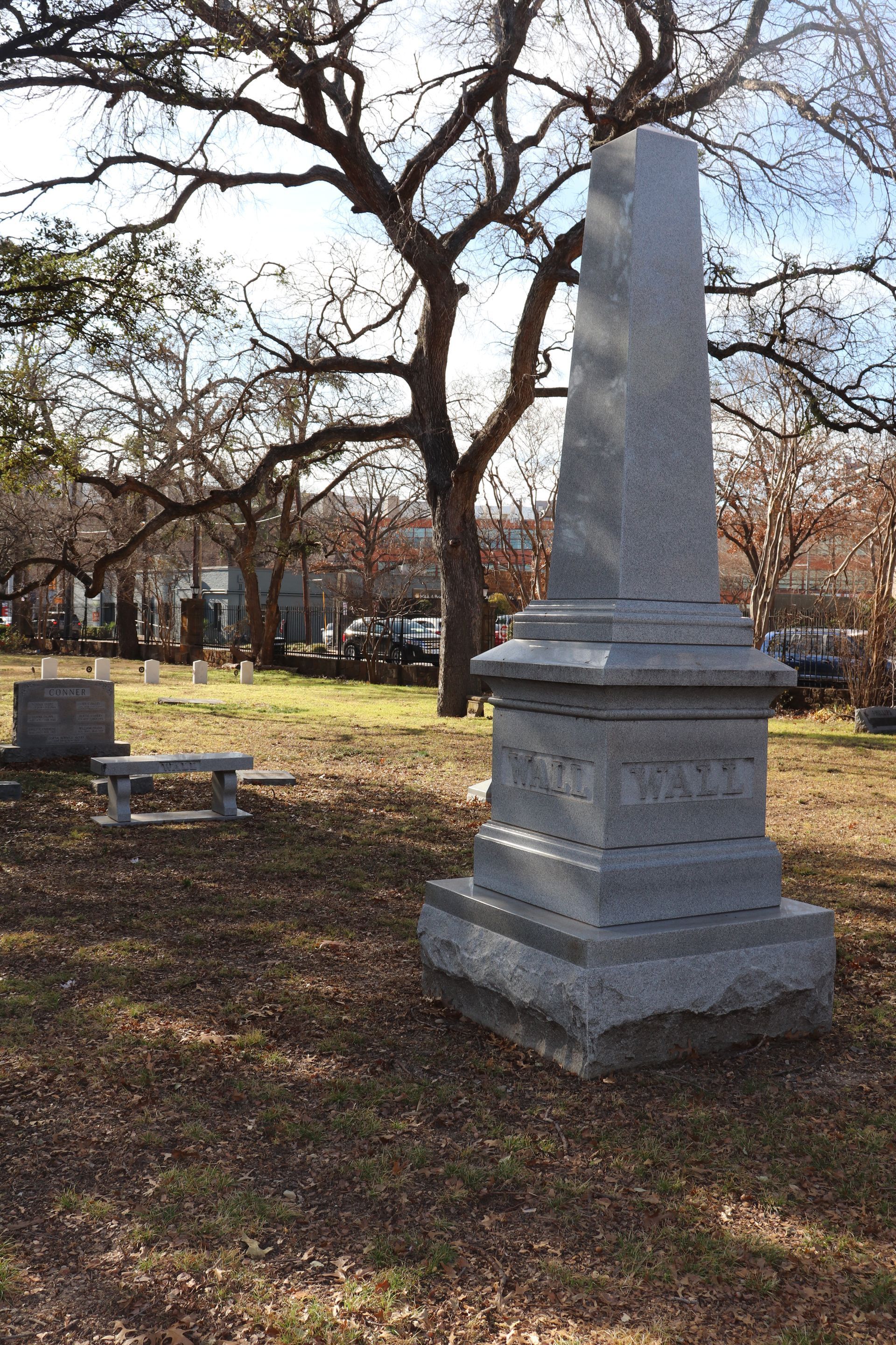 Gray granite obelisk tombstone in a cemetery with a leafless tree in the background.