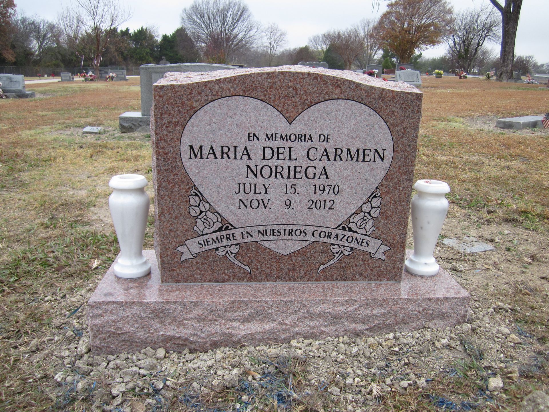 Headstone in a cemetery for Maria del Carmen Noriega, with a heart design and vases on each side.