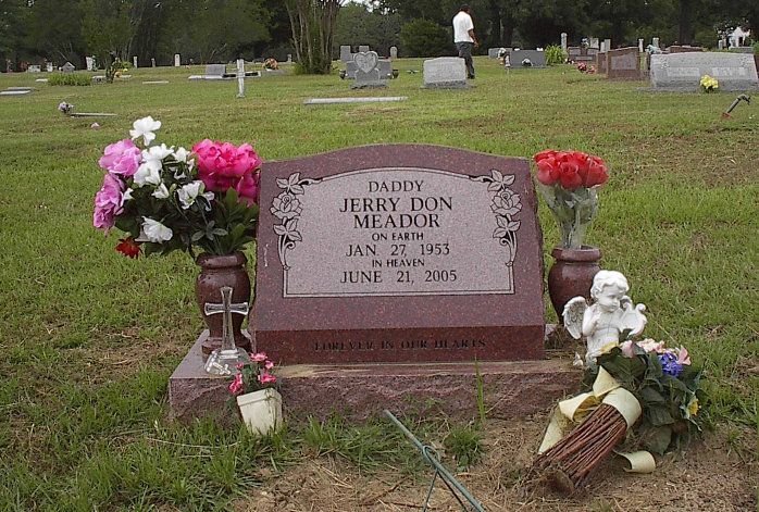 Gravestone in a cemetery with flowers, a small angel statue, and a person in the background.