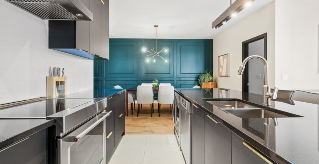 Cuisine moderne avec armoires grises, comptoirs noirs, dosseret bleu turquoise et salle &agrave; manger visible au fond.
