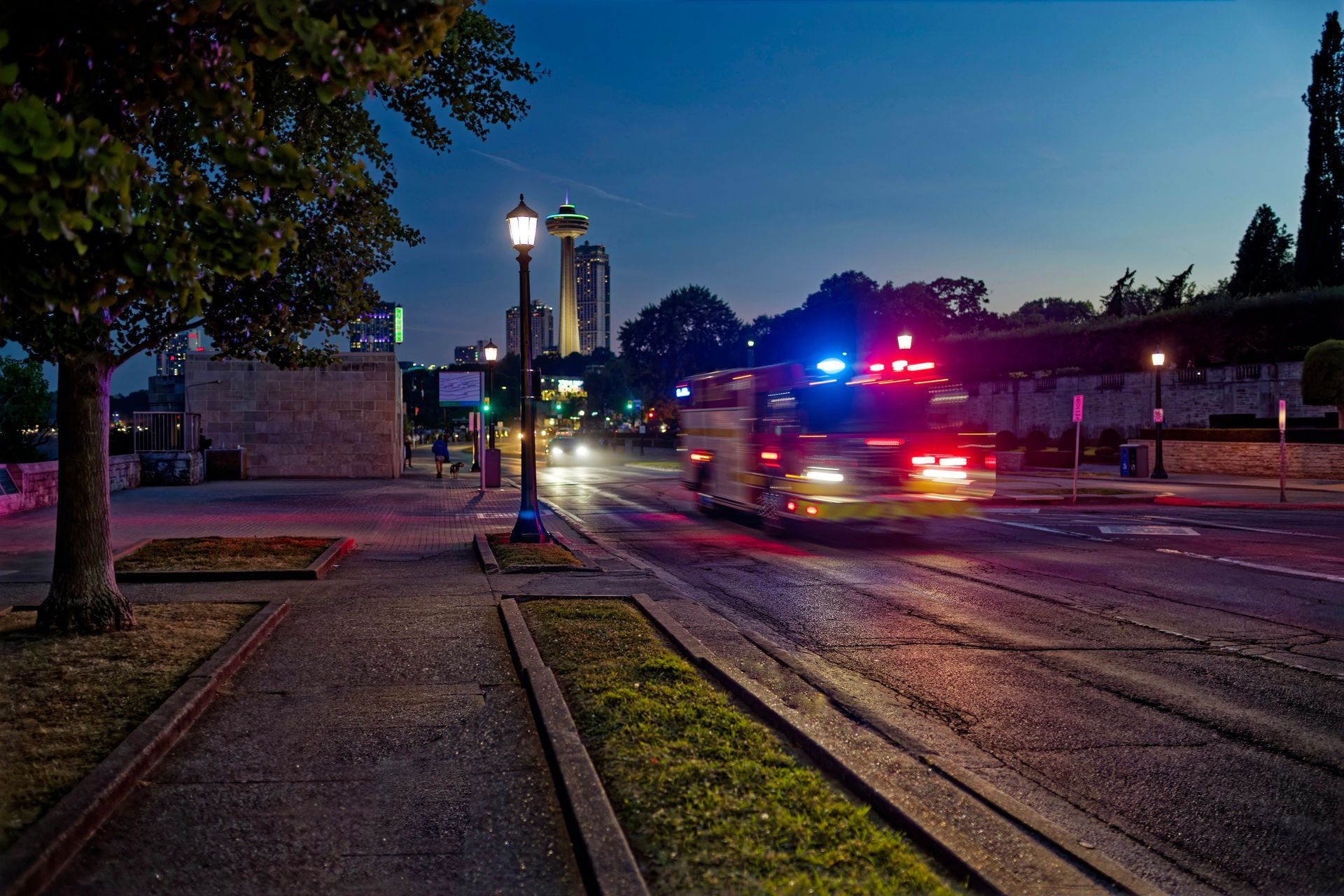 Police car with flashing lights driving on a street at dusk near a sidewalk and trees
