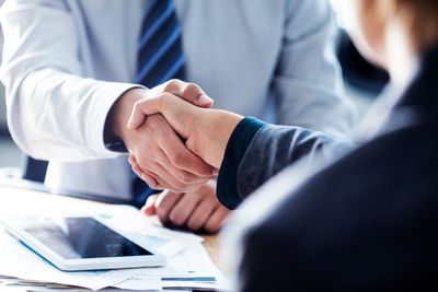 Deux personnes se serrent la main par-dessus un bureau, une tablette et des documents au premier plan.