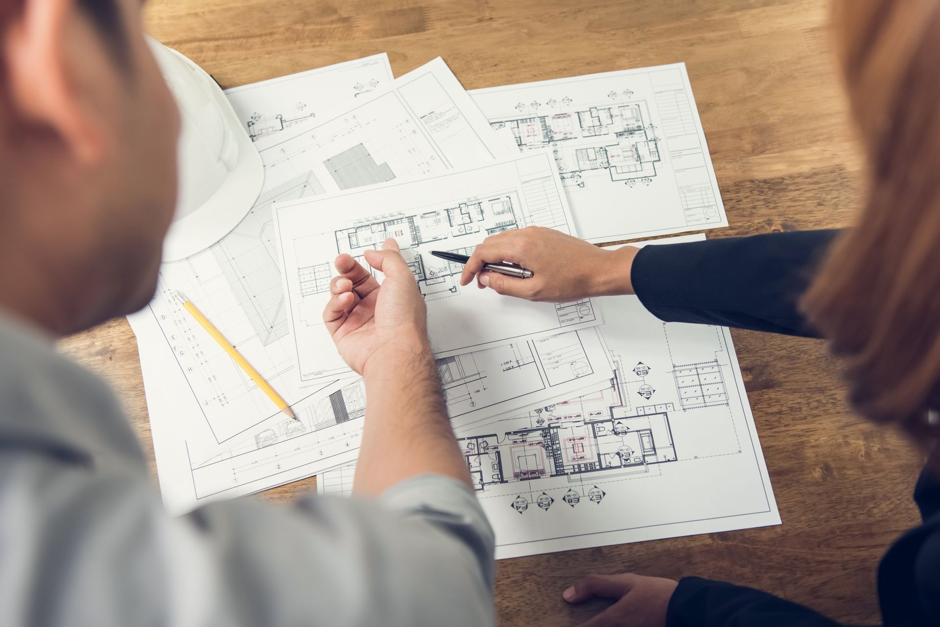 Deux personnes examinent des plans architecturaux sur une table en bois, l'une d'elles désignant un plan.