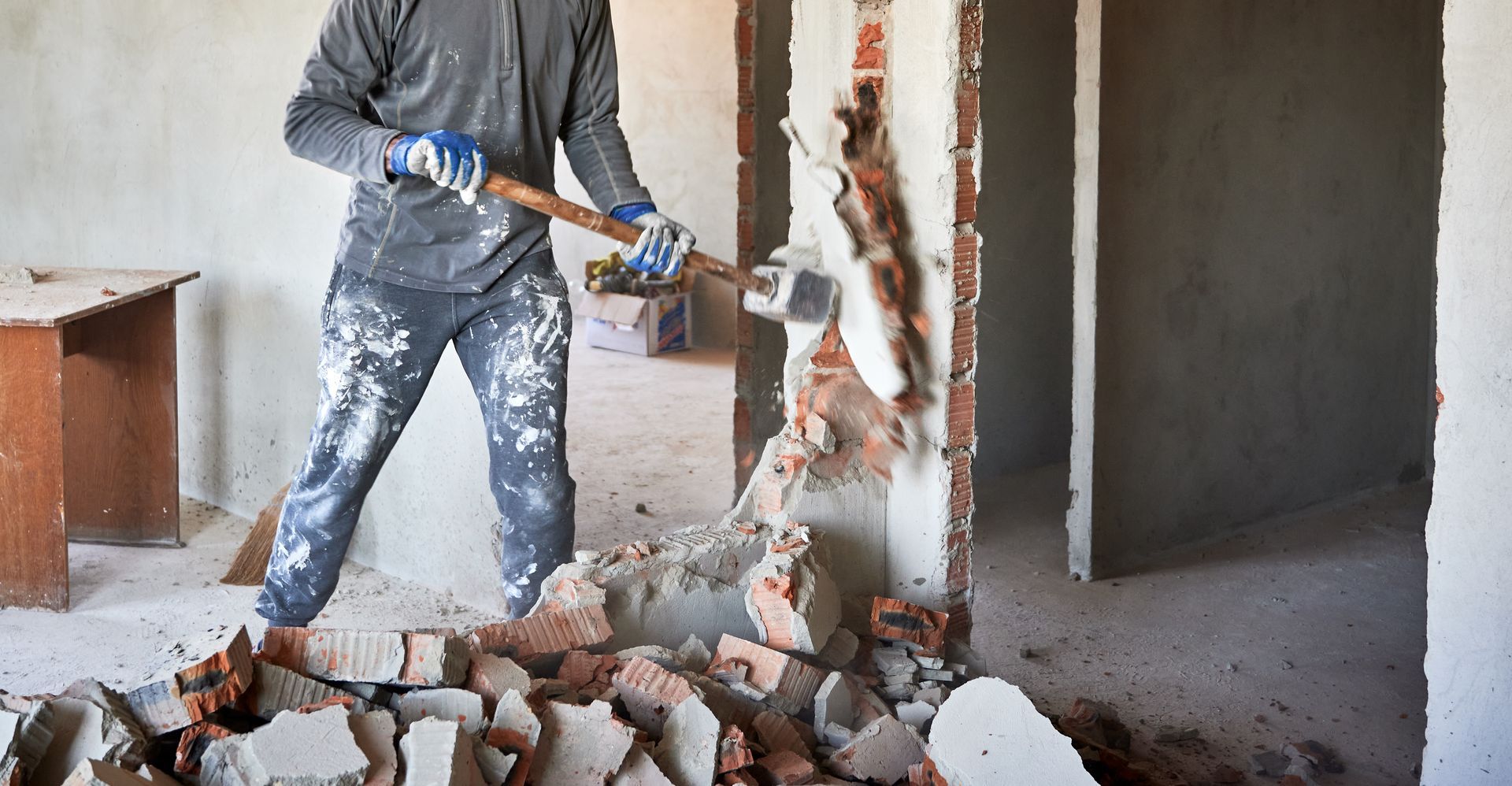 Un ouvrier utilise un marteau-piqueur pour démolir un mur de briques à l'intérieur d'une pièce inachevée.