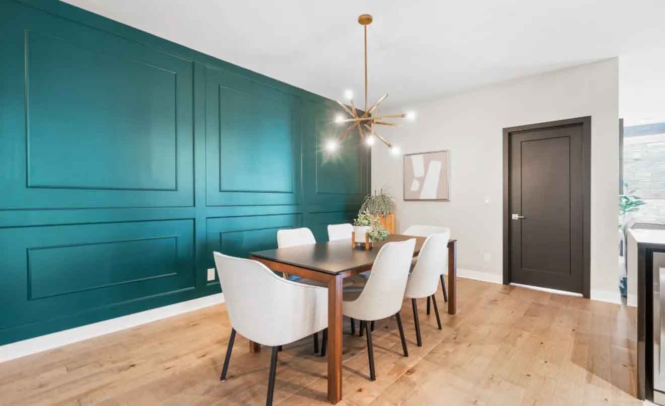 Salle &agrave; manger avec mur turquoise &agrave; panneaux, table en bois, chaises blanches et lustre dor&eacute;.