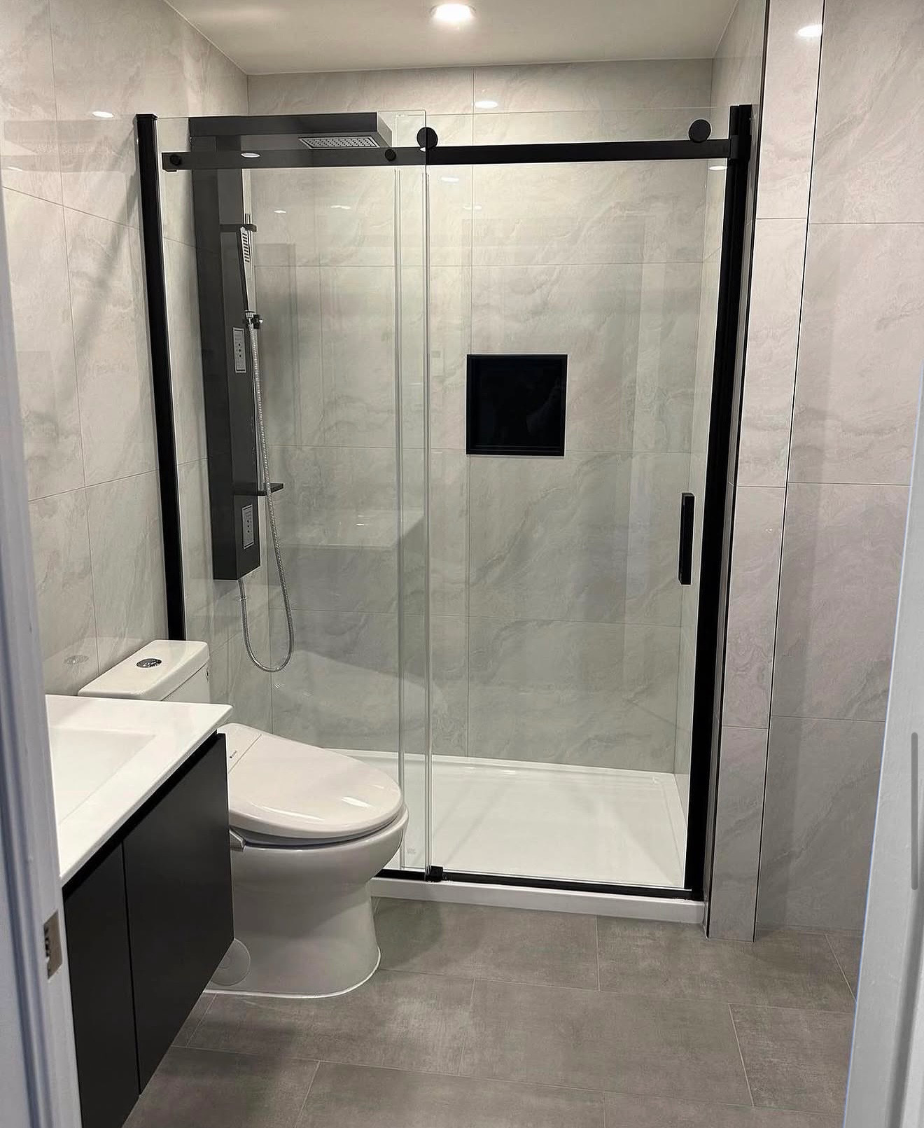 Salle de bains moderne avec cabine de douche en verre, toilettes, lavabo et murs et sol carrelés gris.