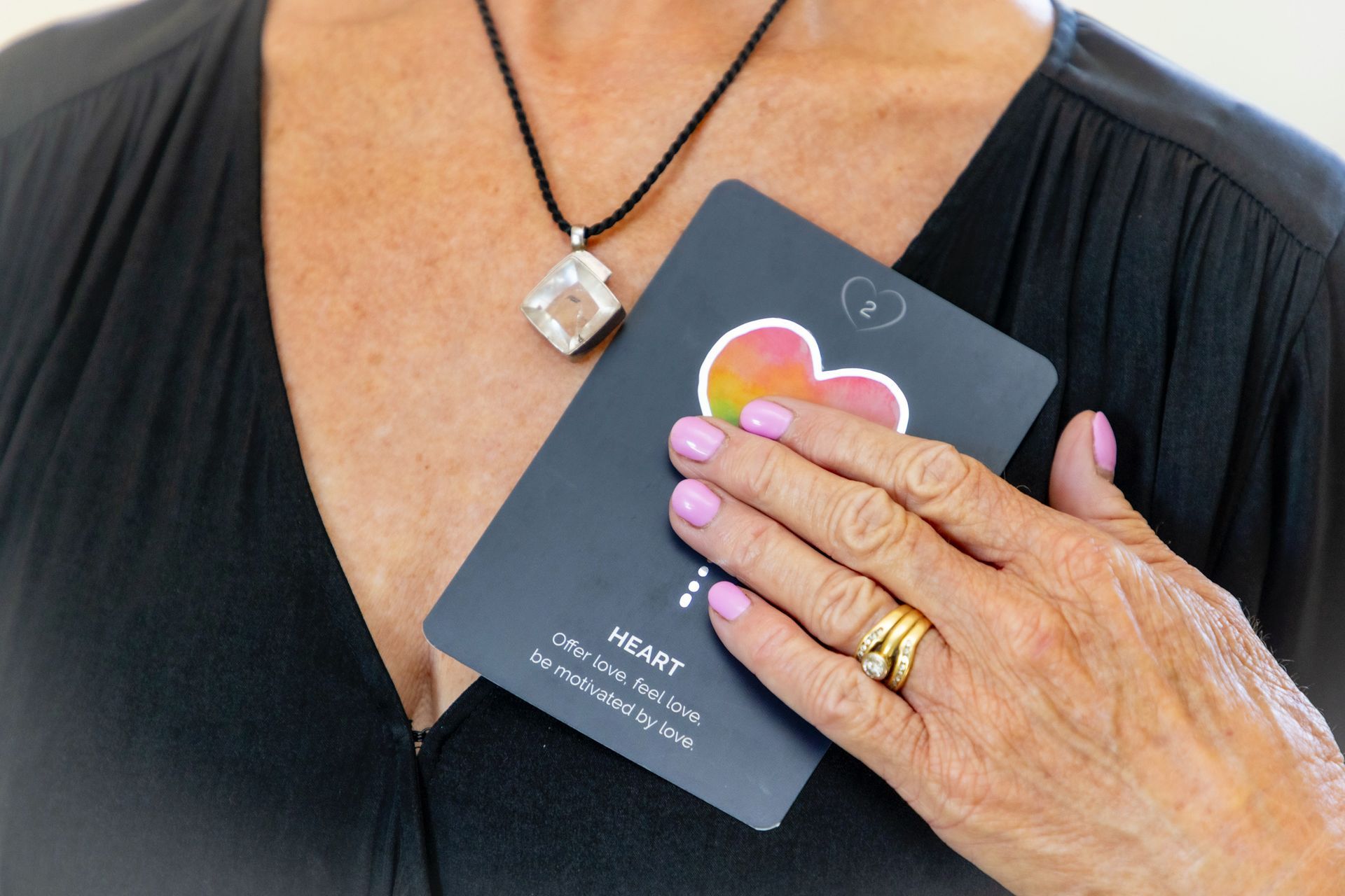 Hand holding a black card with a pink heart over a black blouse and necklace — Soulfully Happy In Springvale, NSW
