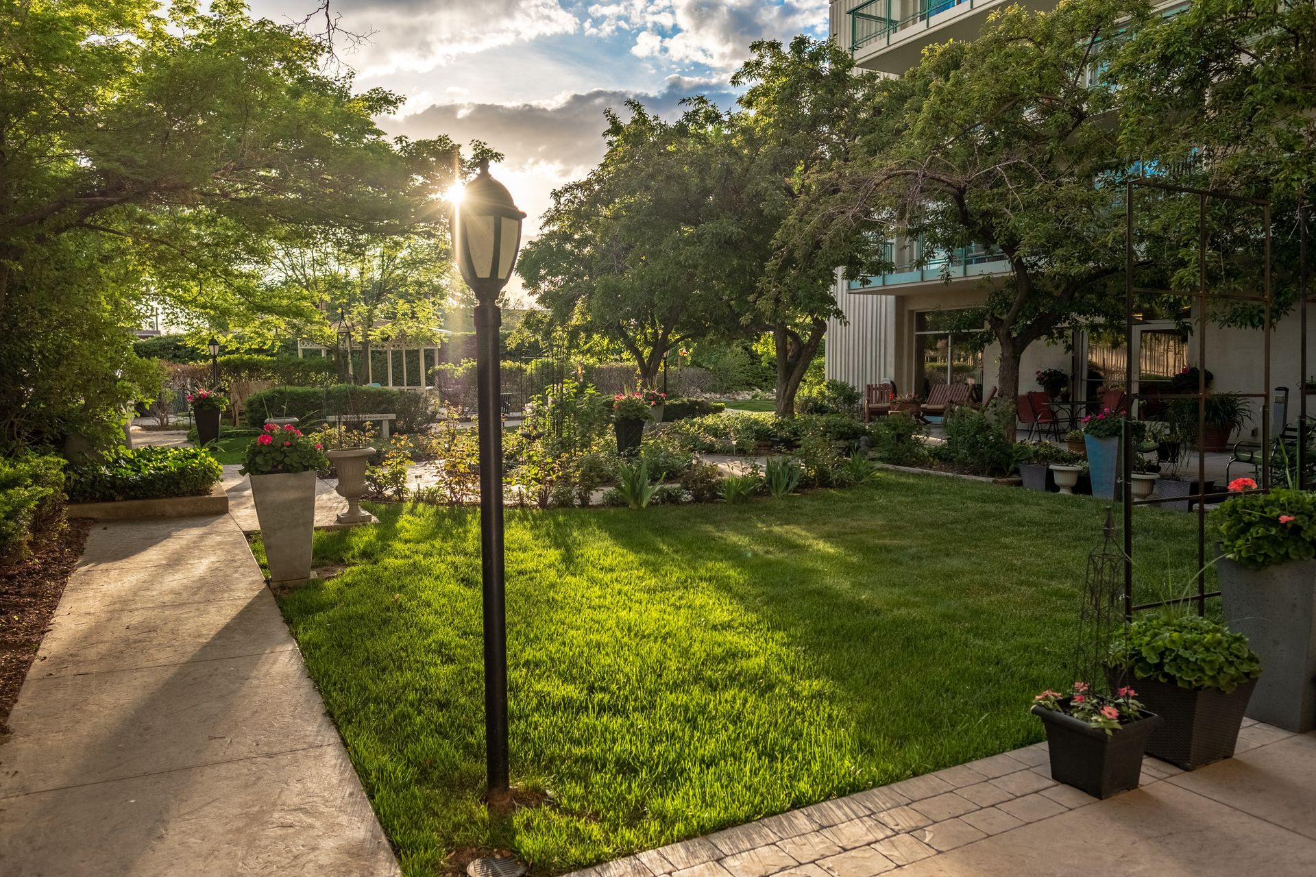 Sunny garden courtyard with lush green lawn, stone paths, potted plants, and patio seating around a central lamp.