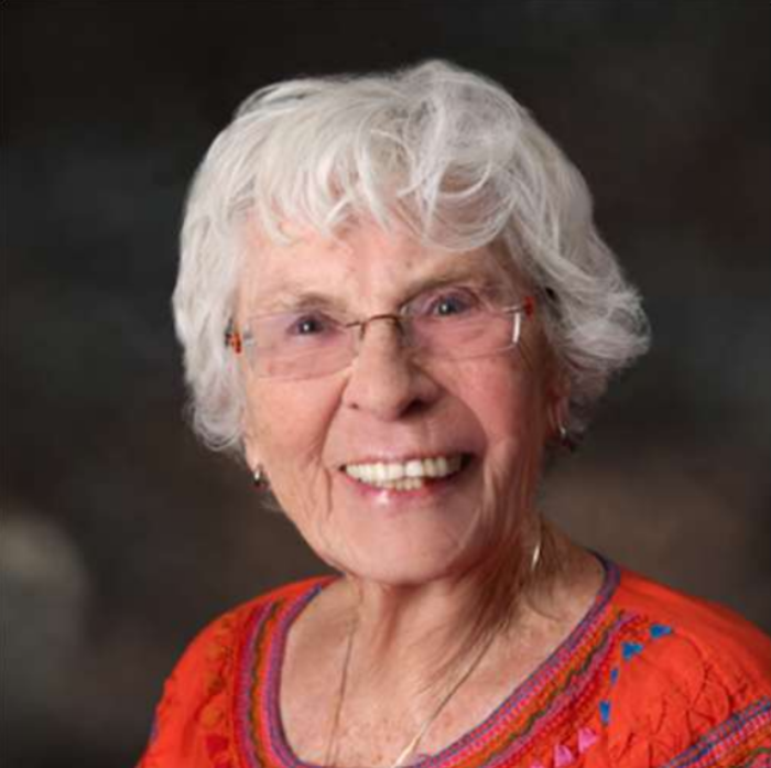 Smiling woman with white hair and glasses wearing a red-orange patterned top against a dark background
